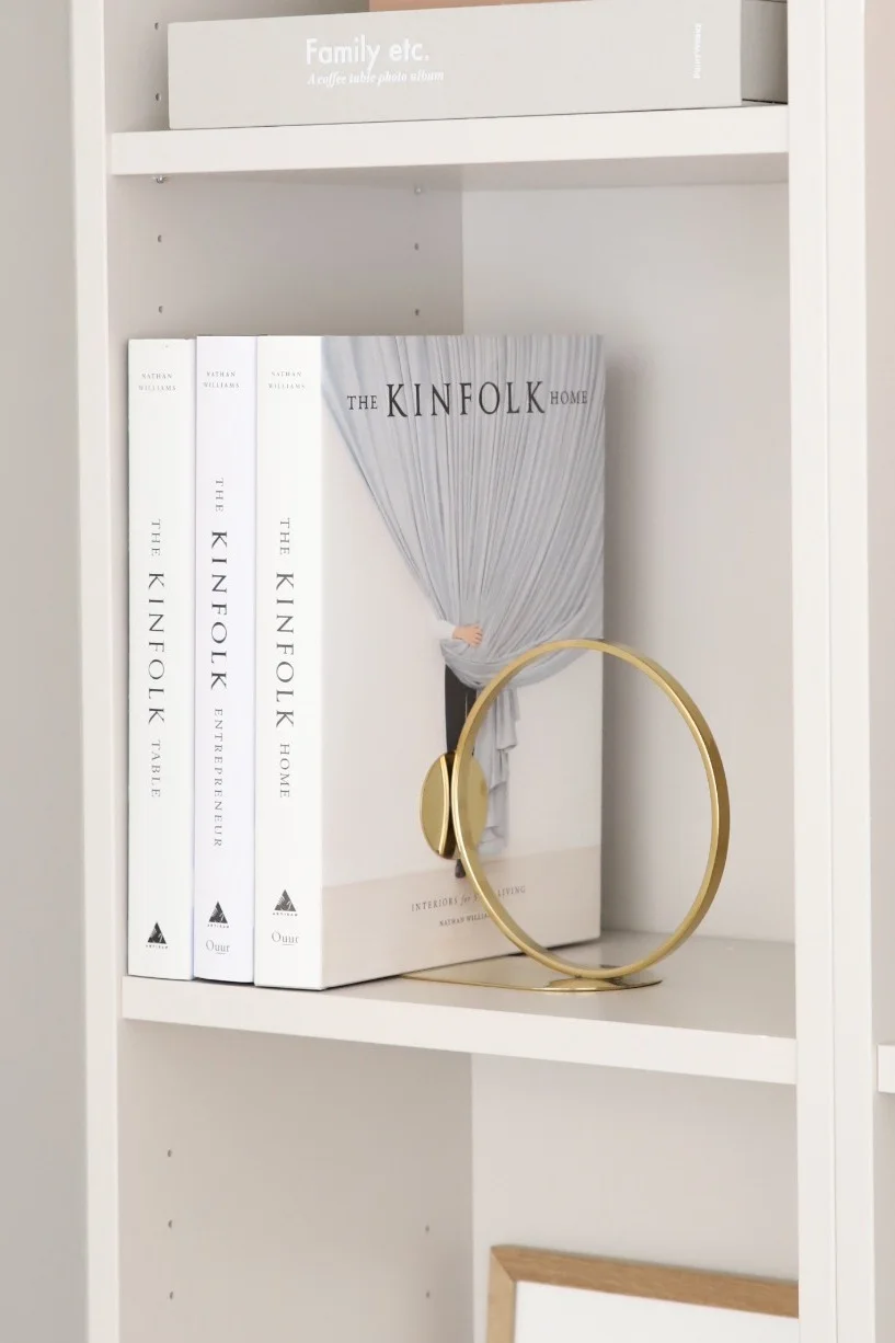 A white bookshelf with multiple Kinfolk books, including 'The Kinfolk Home' standing cover-out with a modern gold circular bookend.
