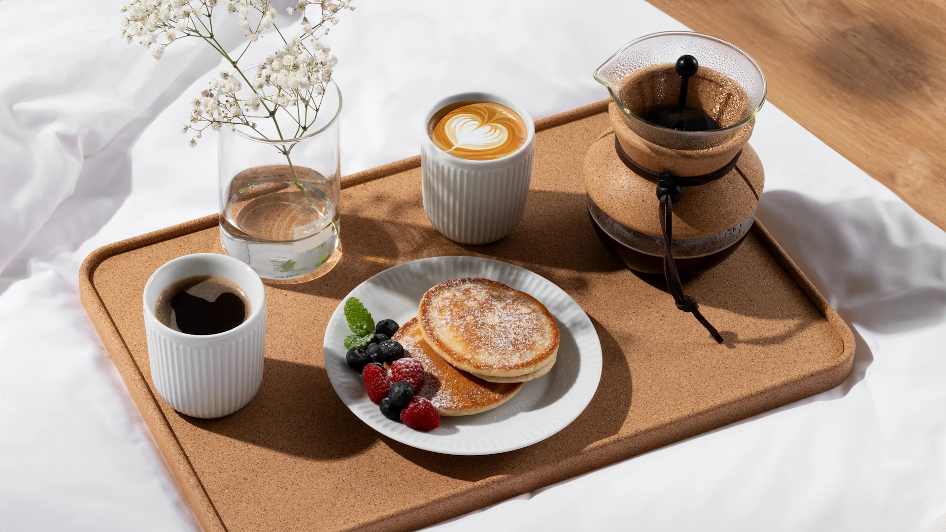 Breakfast in bed: pancakes with berries and powdered sugar, two coffees, and flowers on a cork tray.