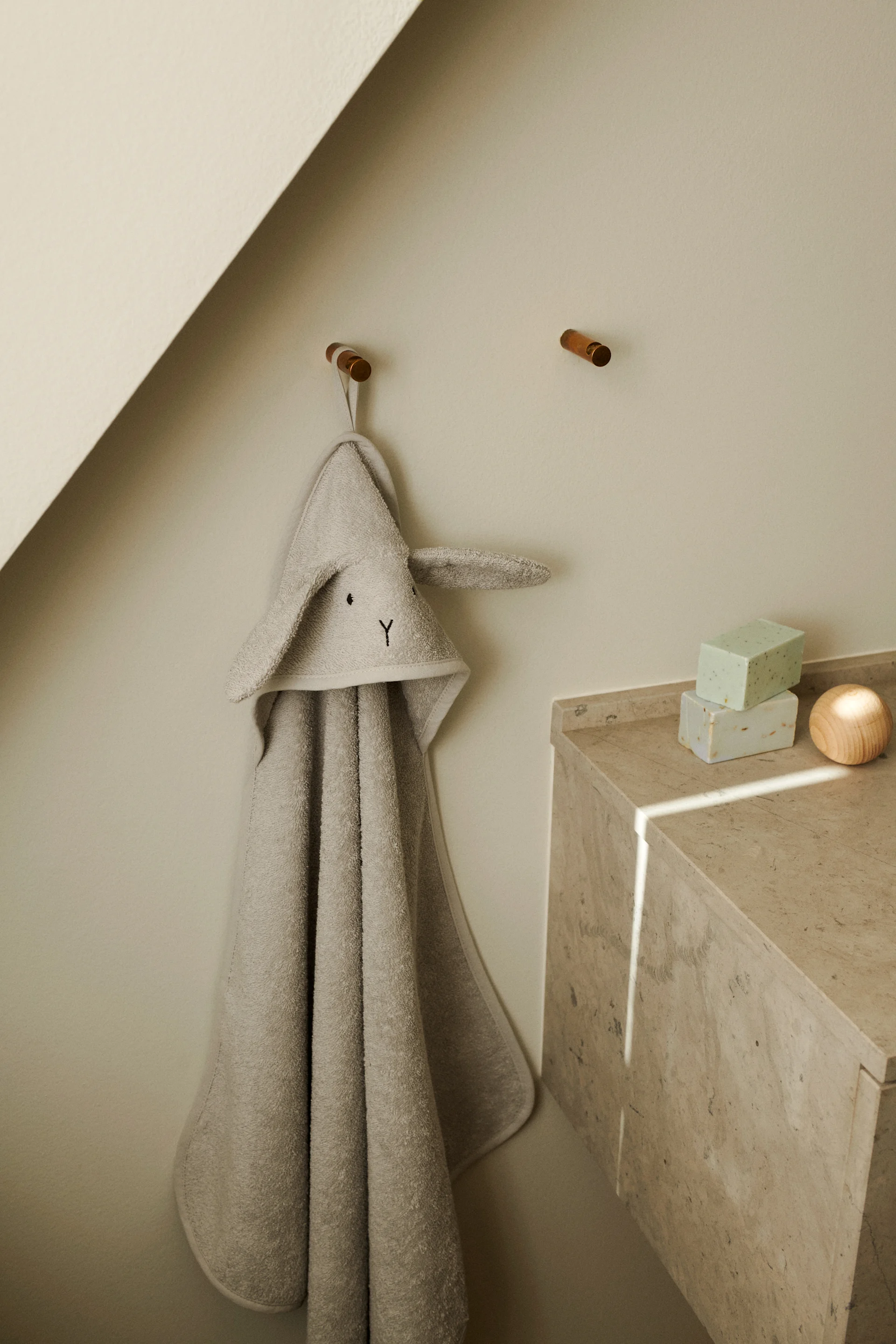 A light gray hooded towel with bunny ears hangs on a wooden peg, next to a marble counter with two green soaps and a wooden ball.