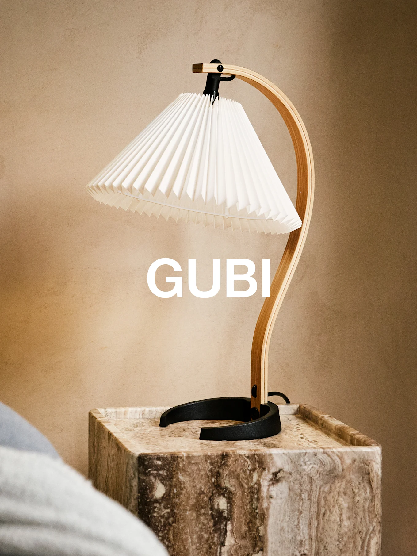 GUBI table lamp with a white pleated shade and curved wooden arm on a marble side table, with GUBI text behind it.