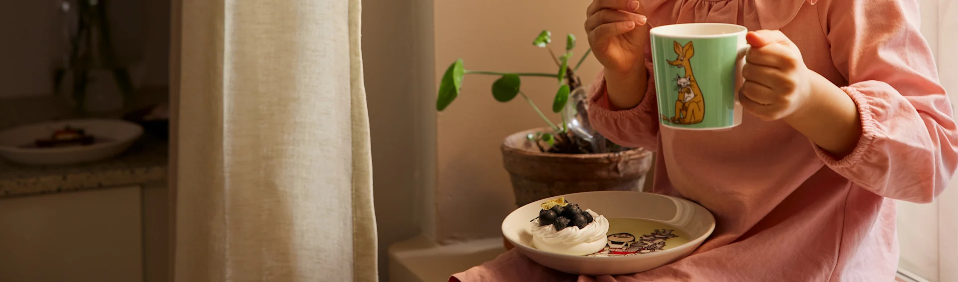 Person in pink shirt holding a green mug and a bowl with meringue and blueberries.