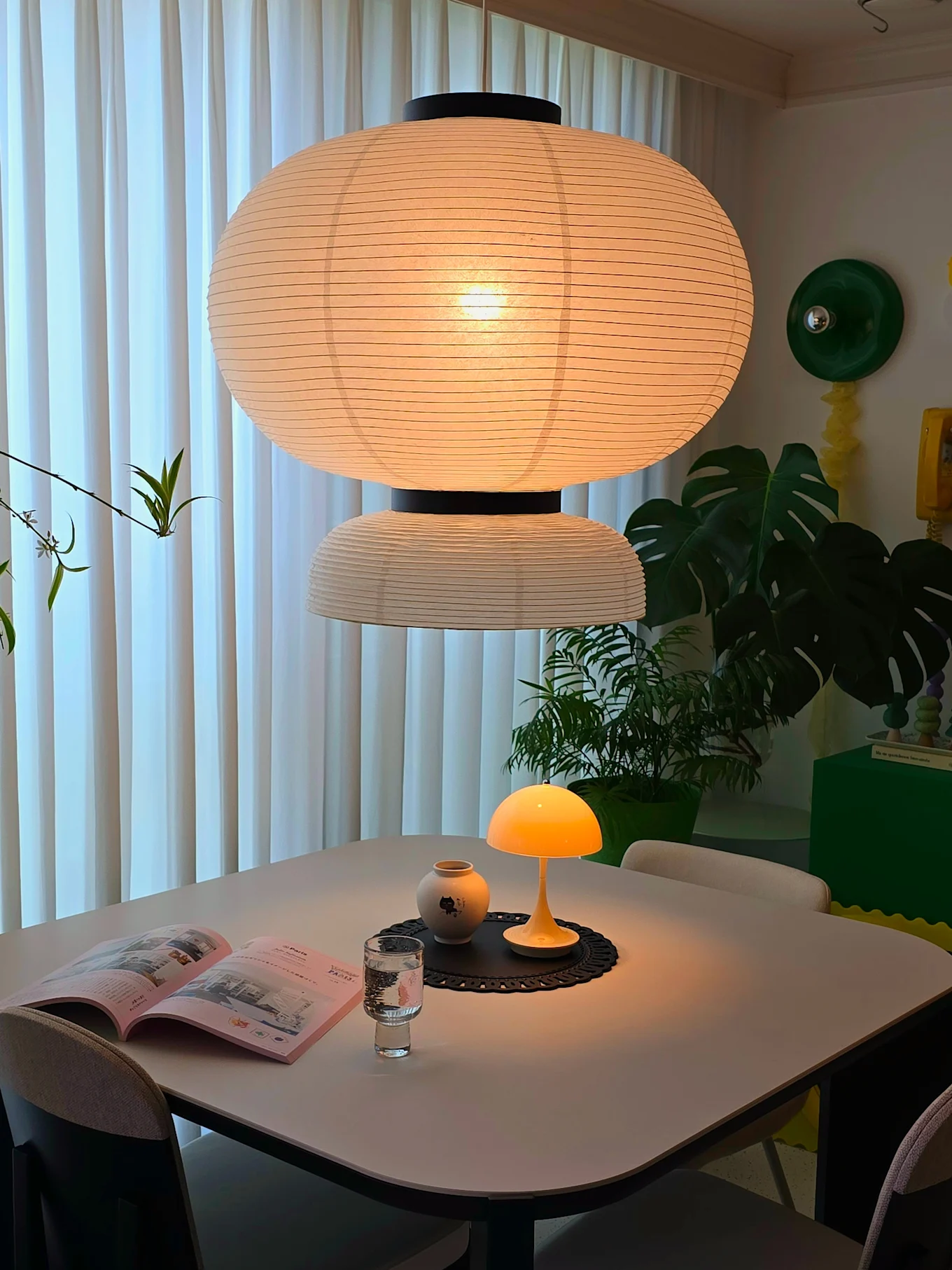 A cozy dining area featuring a large paper lantern pendant light and a glowing yellow mushroom lamp on the table with a book and vase.