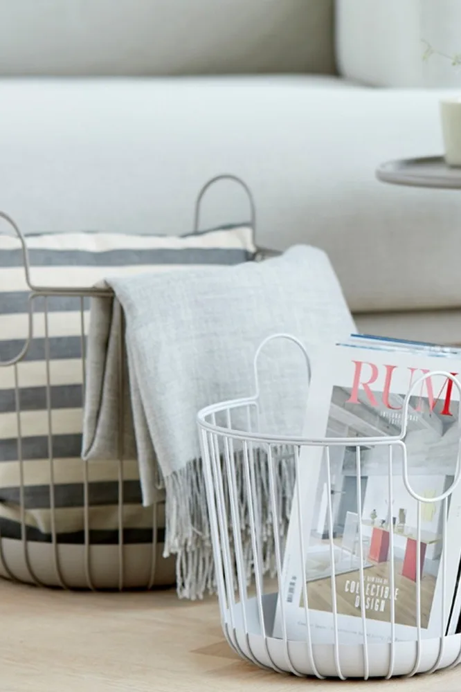 Two round wire baskets, one gray with a striped pillow and throw, one white with magazines, on a light wooden floor.