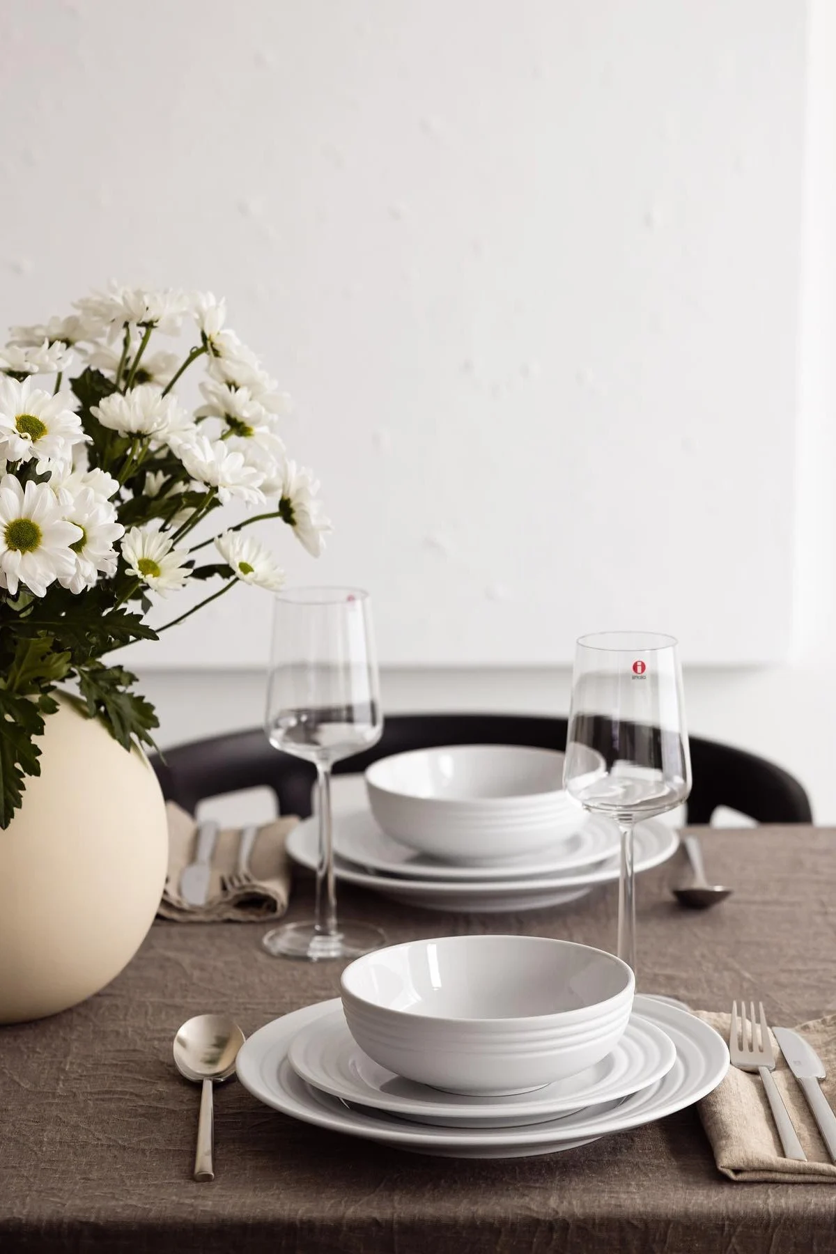 A dining table set for two with white plates and bowls, clear wine glasses, and a vase of white flowers on a brown tablecloth.