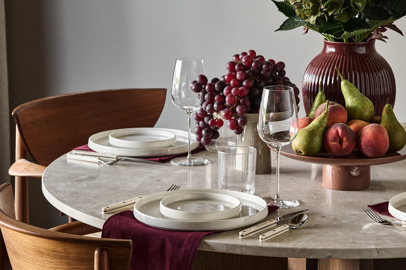 Table with plates, cutlery, glasses, red grapes, green pears, peaches, and a red ribbed vase with foliage.