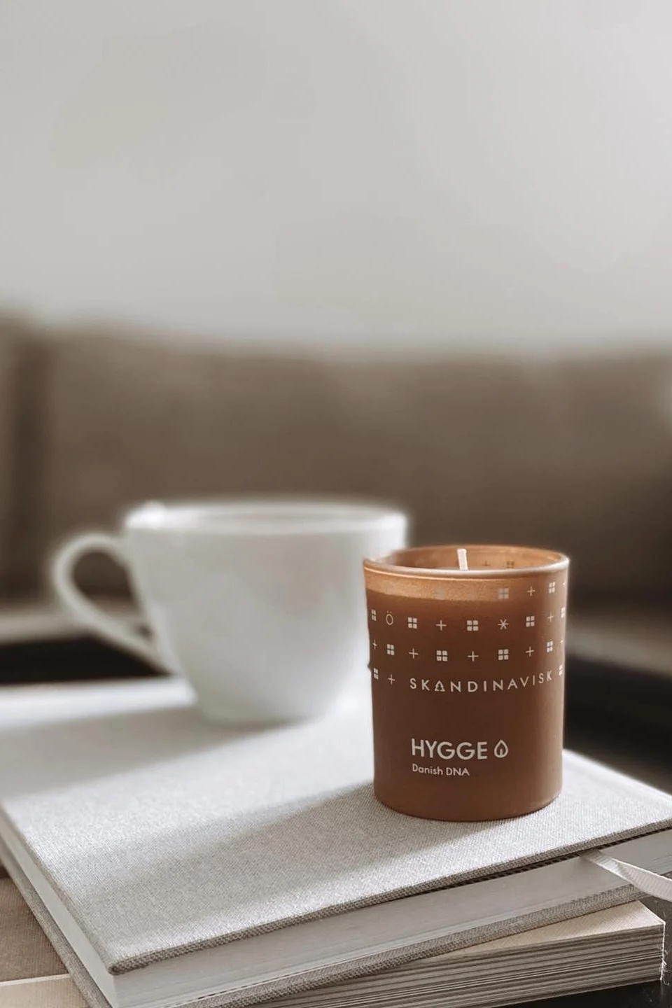 A brown candle with 'HYGGE SKANDINAVISK' text sits on a book, with a white mug blurred in the background.