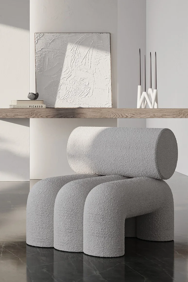 Light grey bouclé chair with cylindrical back and three curved legs, in a modern room with artwork and candles.