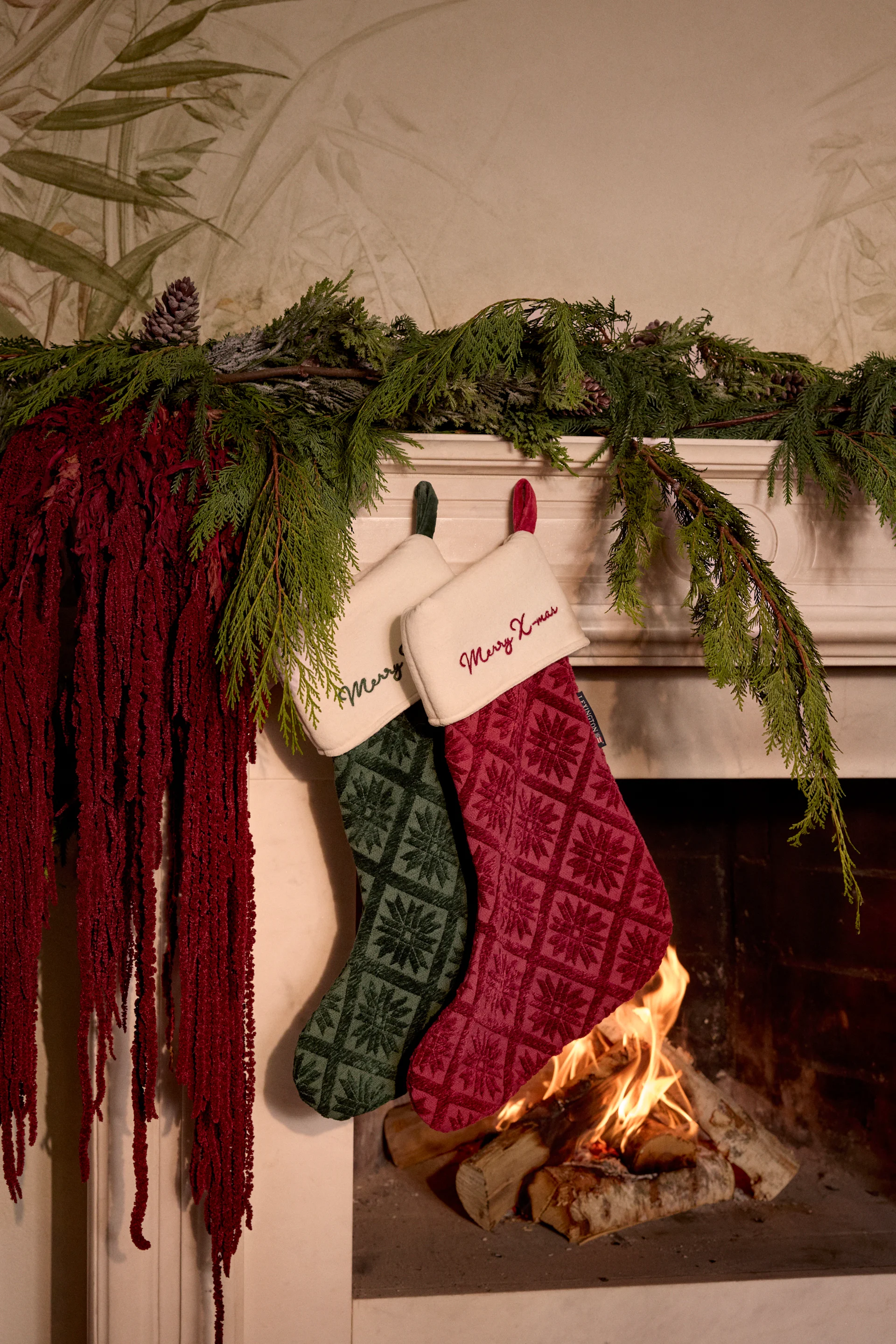 Two patterned Christmas stockings, one green and one red, hanging above a lit fireplace, decorated with evergreen garland and red foliage.