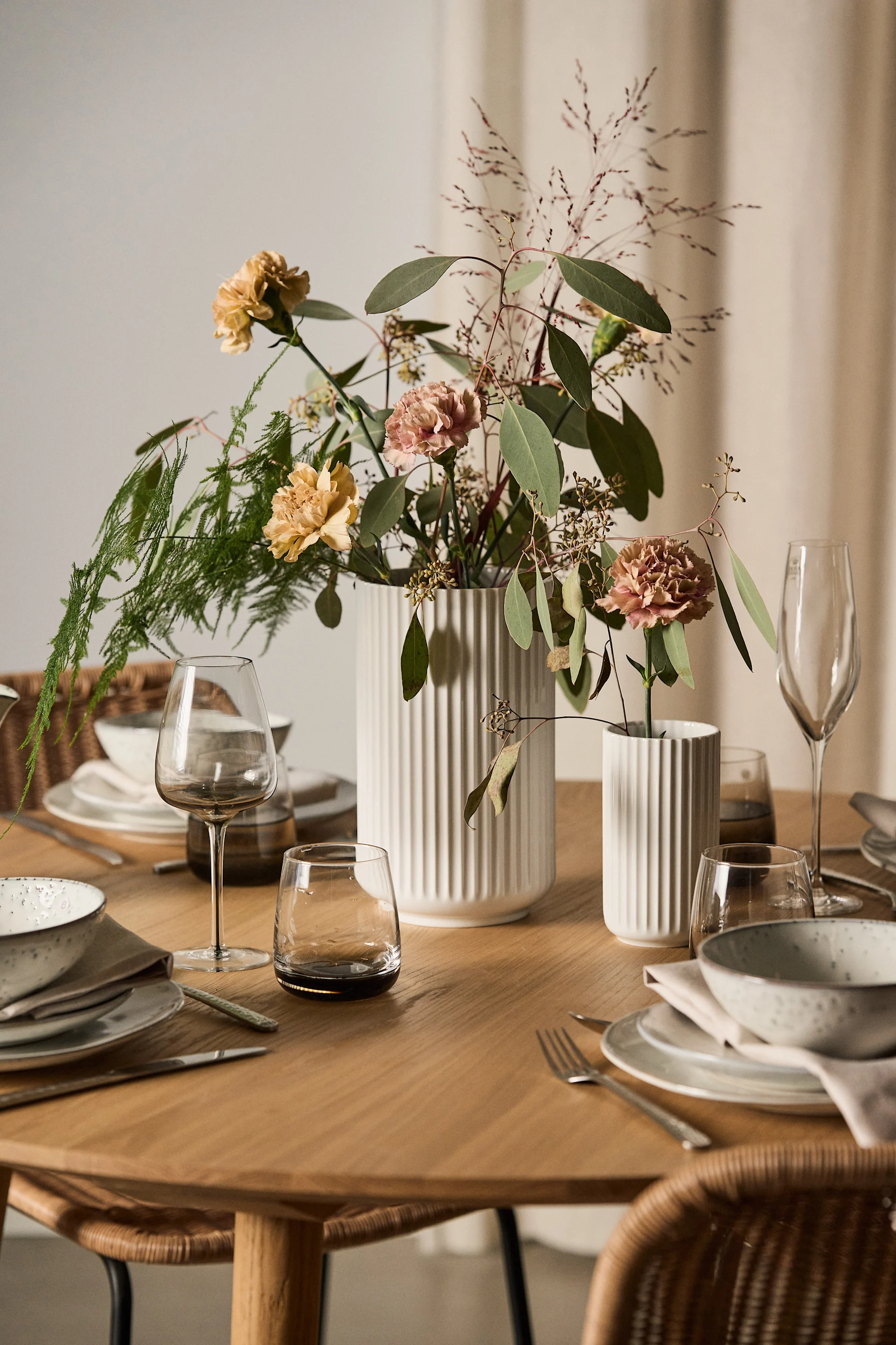Elegantly set dining table with white and speckled dinnerware, clear and smoked glassware, and a floral centerpiece in white vases.