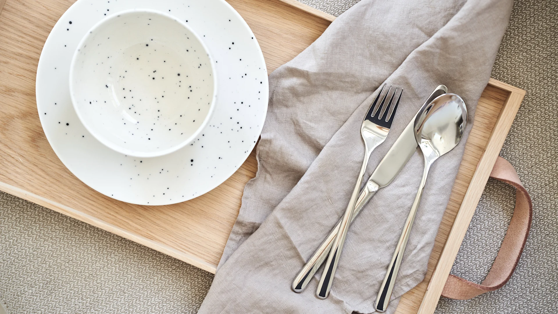 Overhead view of a wooden tray with a white speckled bowl and plate, a neutral linen napkin, and silver cutlery.