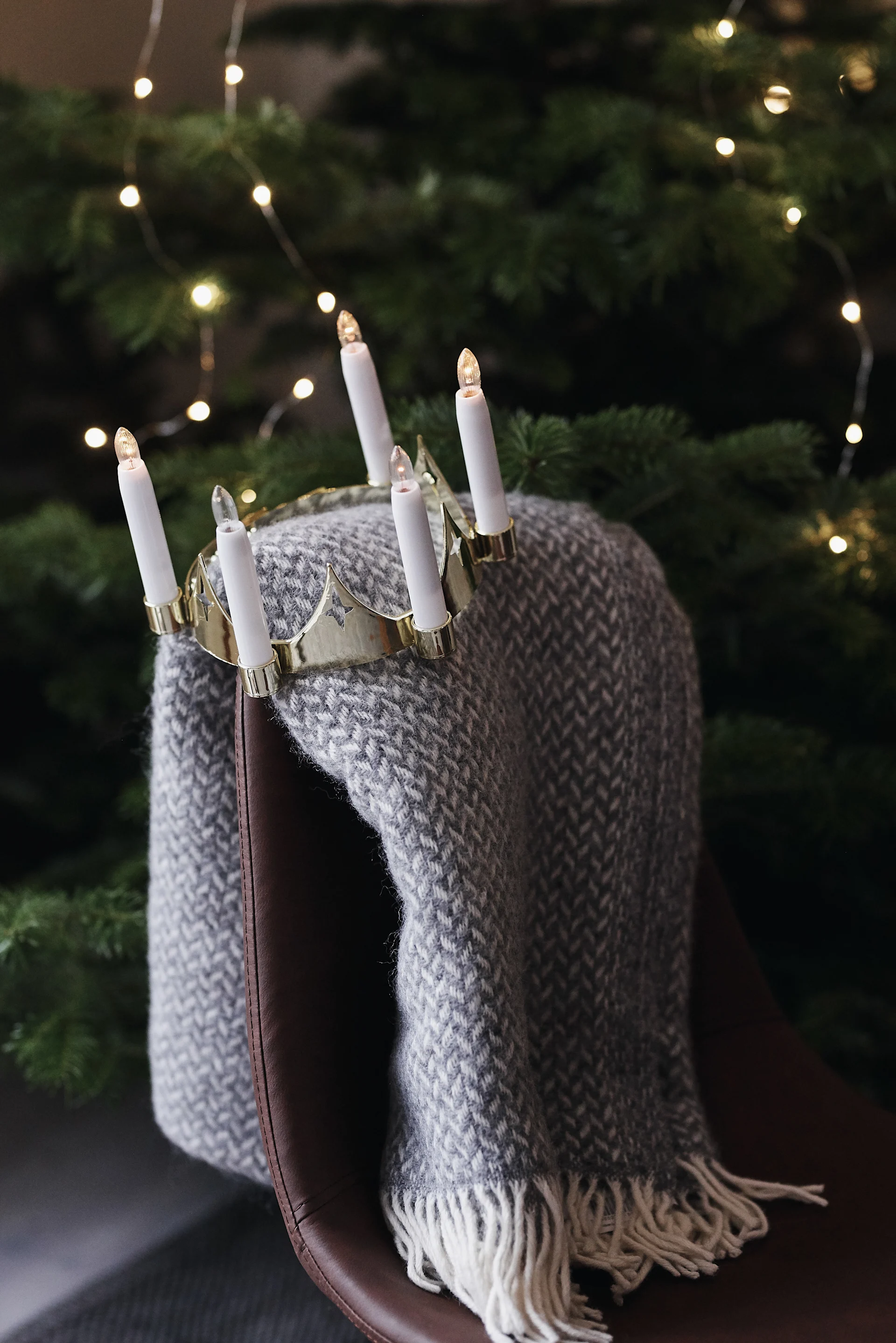 Gold crown-shaped candelabra with five electric candles on a grey herringbone throw draped over a brown leather chair, with Christmas tree lights.