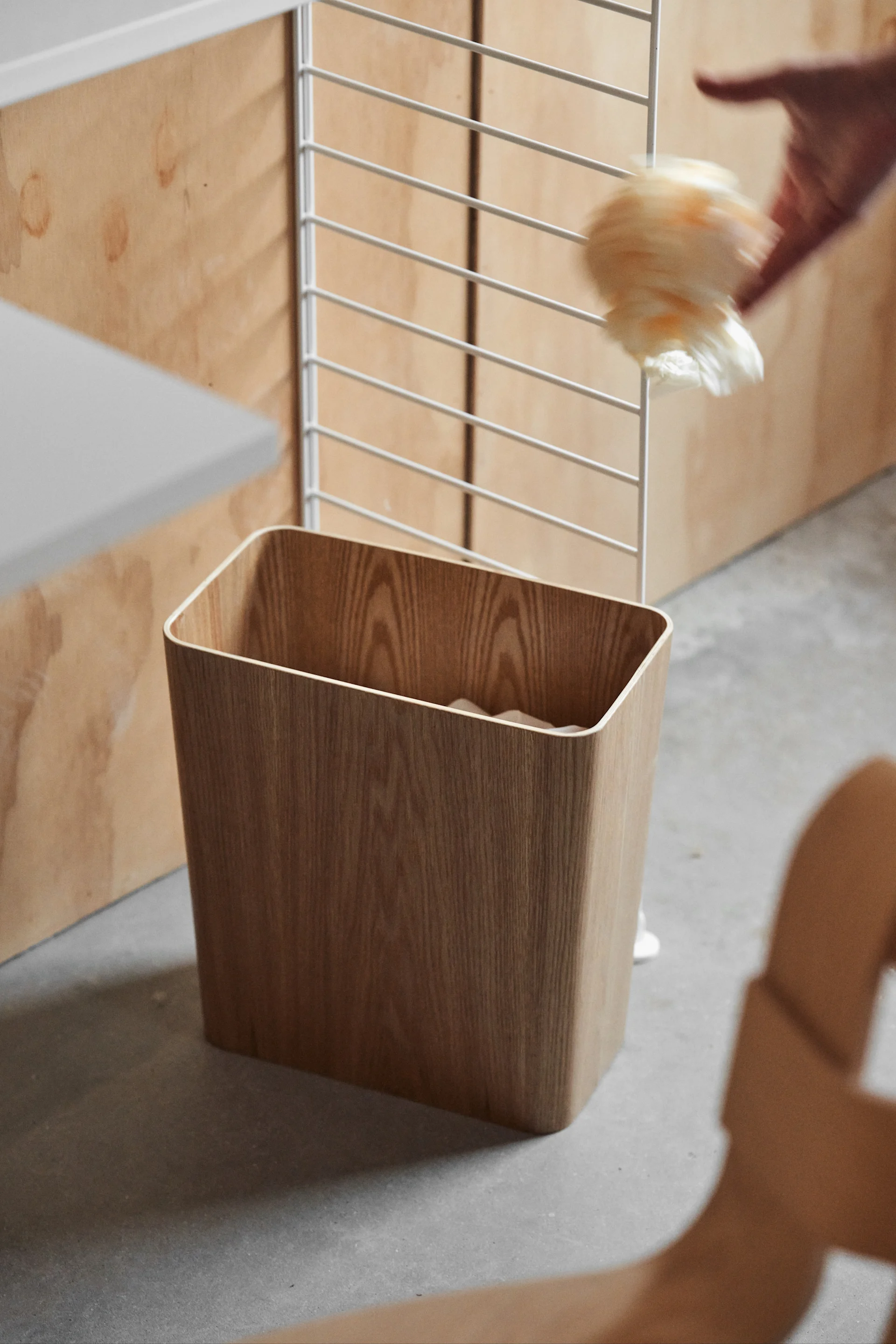 A hand drops a fluffy item into a light wood waste bin, next to a wood-paneled wall and metal shelf.