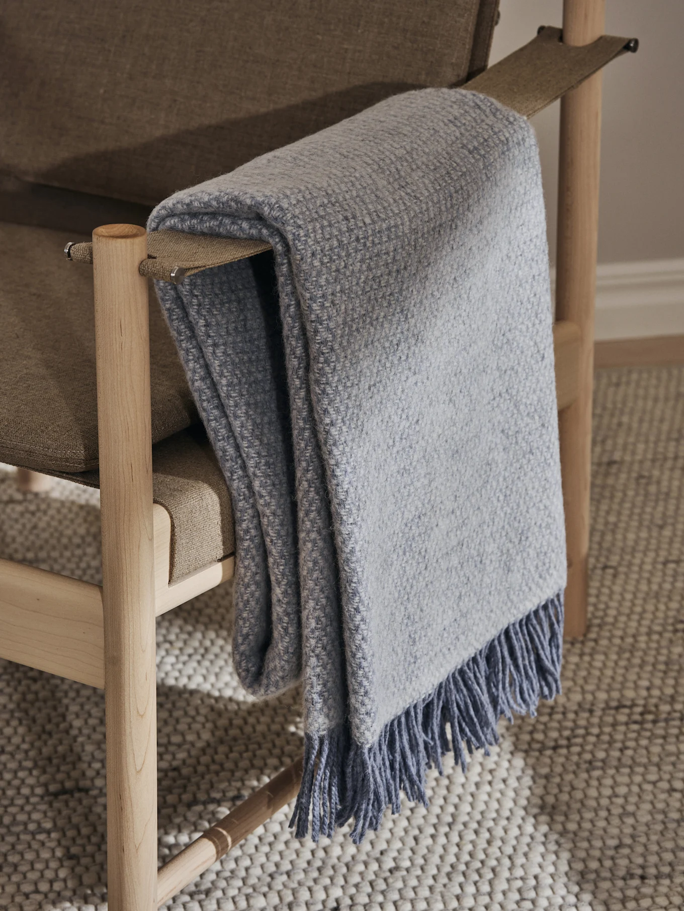 Light blue woven blanket with fringes draped over a light brown upholstered chair with a light wooden frame, on a textured rug.