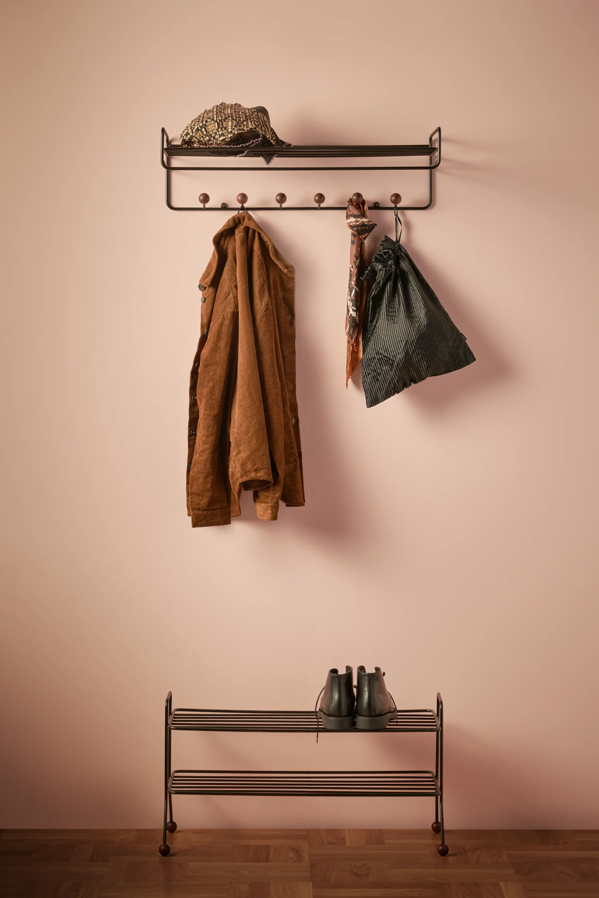 Coat rack with a brown corduroy jacket, patterned hat, scarf, and dark drawstring bag, above a shoe rack with black boots, against a light pink wall.