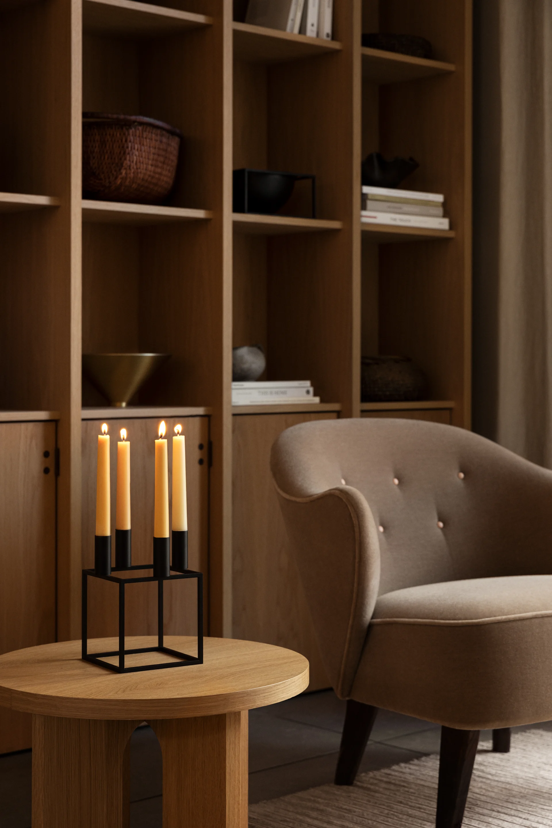 Four lit yellow candles in a black metal holder on a round wooden table, next to a beige armchair and a wooden bookshelf.