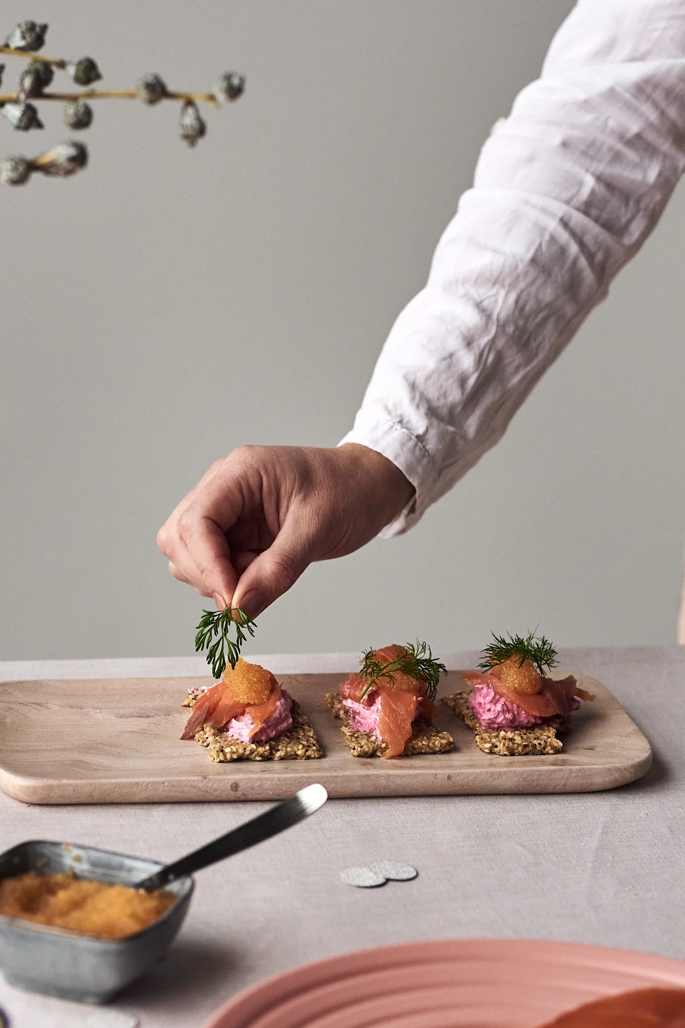 A hand garnishes crispbread appetizers topped with pink spread, smoked salmon, and orange caviar with fresh dill on a wooden board.