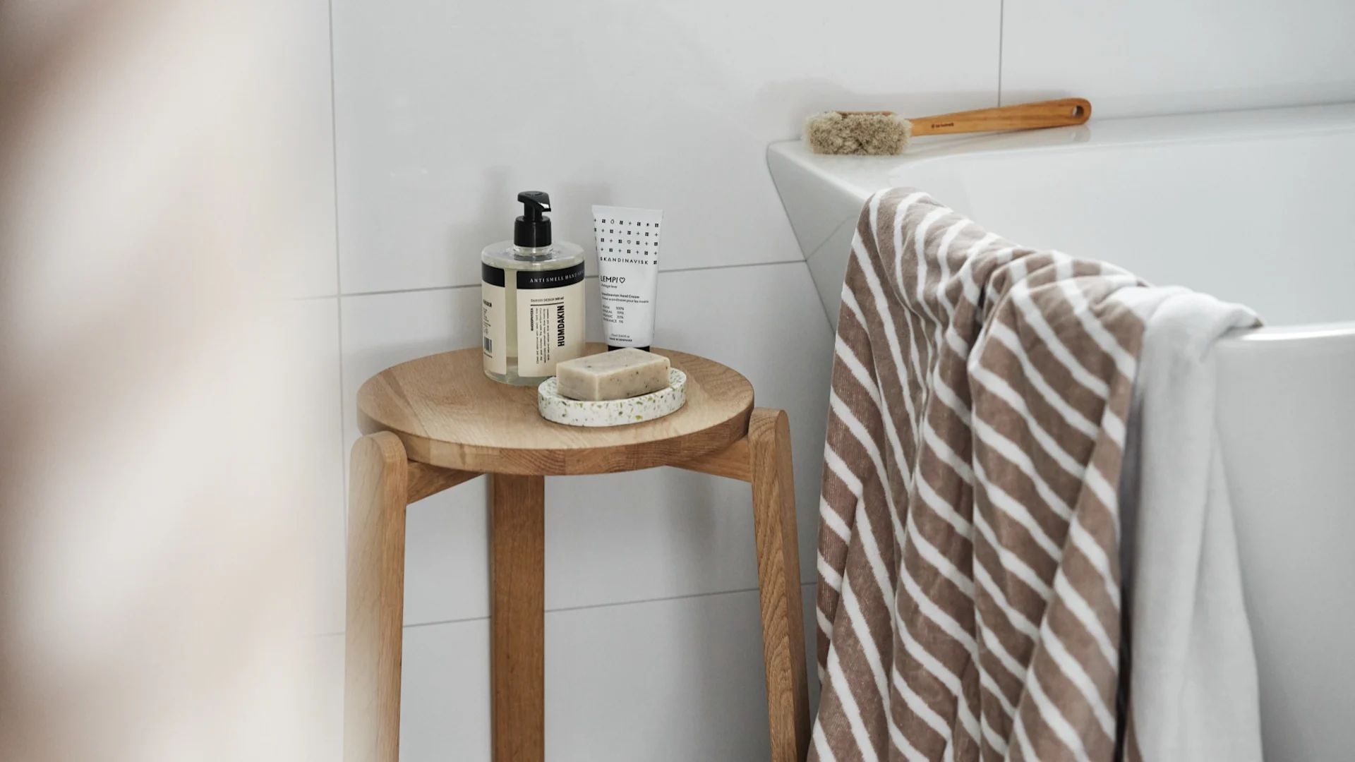 Bathroom scene with wooden stool holding soap pump, bar soap, hand cream. Striped towel on white tub.