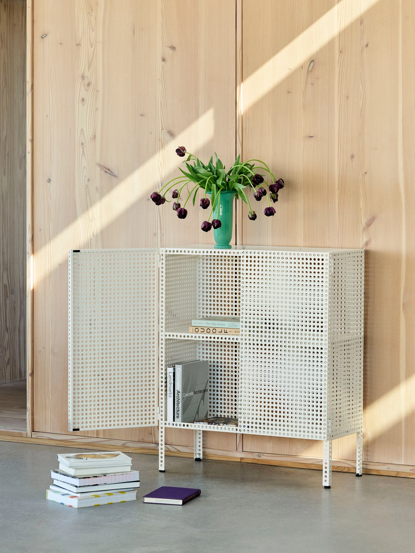 White perforated metal cabinet with open doors, green vase with purple tulips on top, books inside and on the floor.