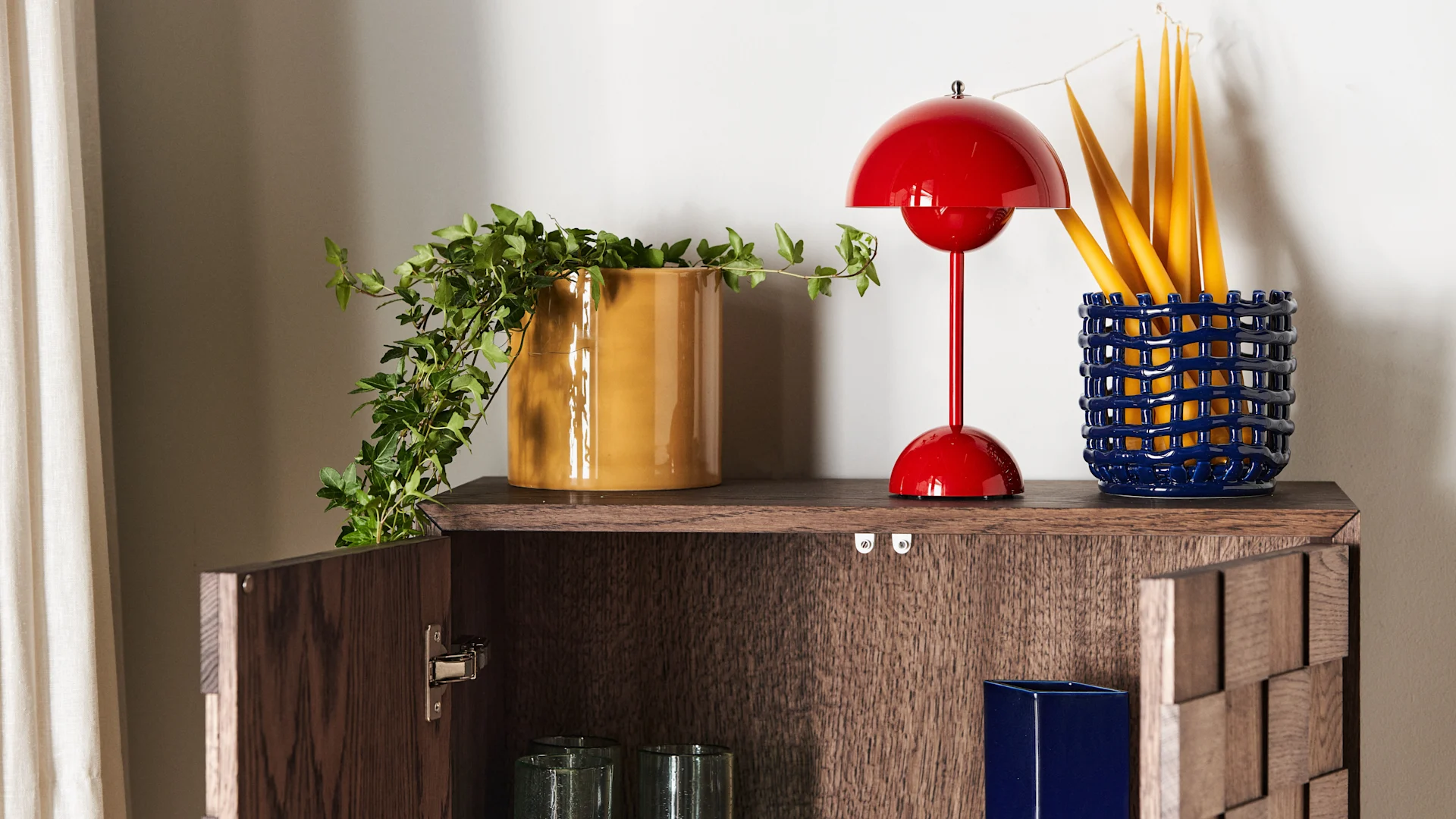 A dark wood cabinet with a red dome lamp, an ivy plant in a yellow pot, and yellow candles in a blue woven pot on top.