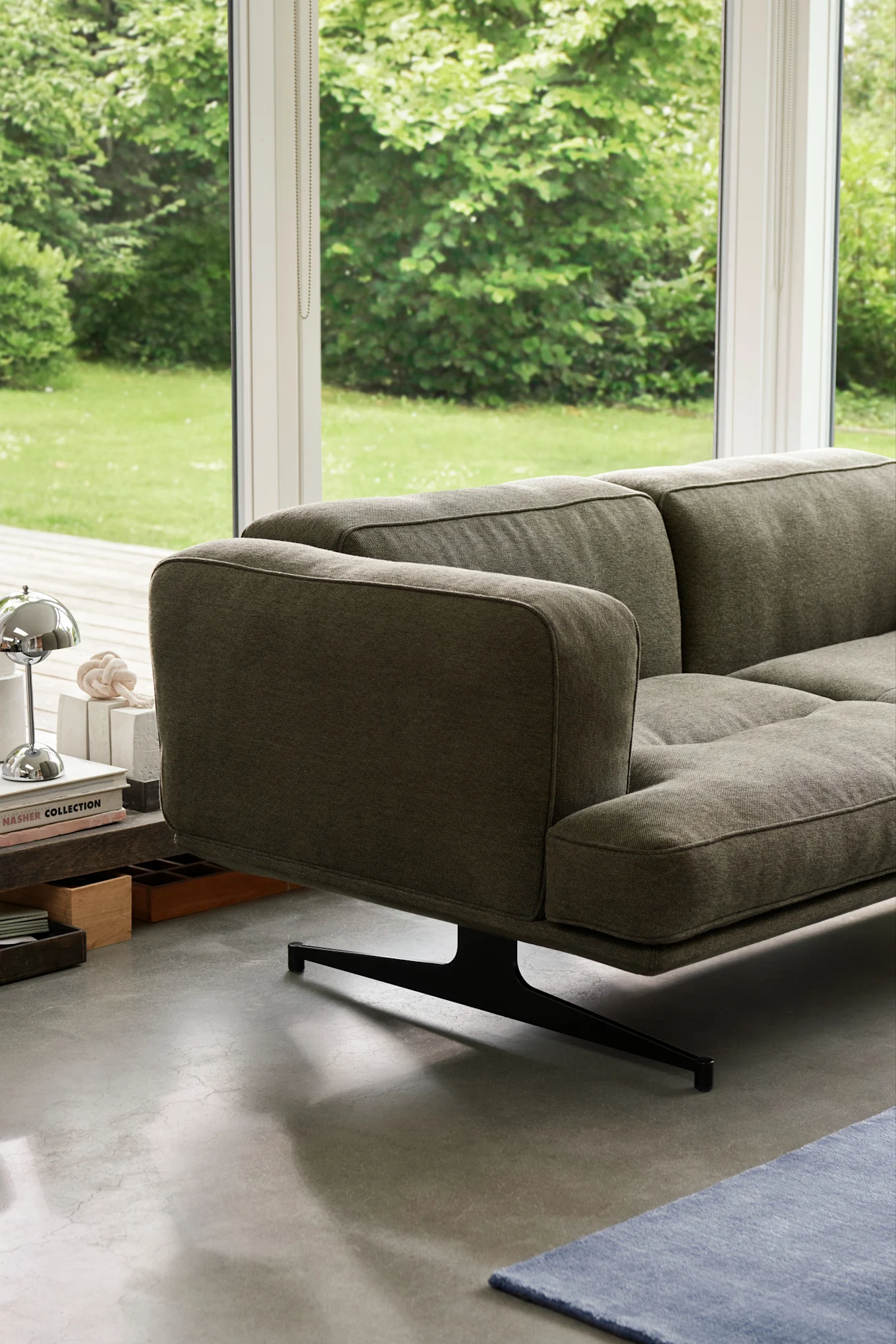 Modern grey fabric sofa with a black sculptural base, a side table with books, and a view of a lush green garden.