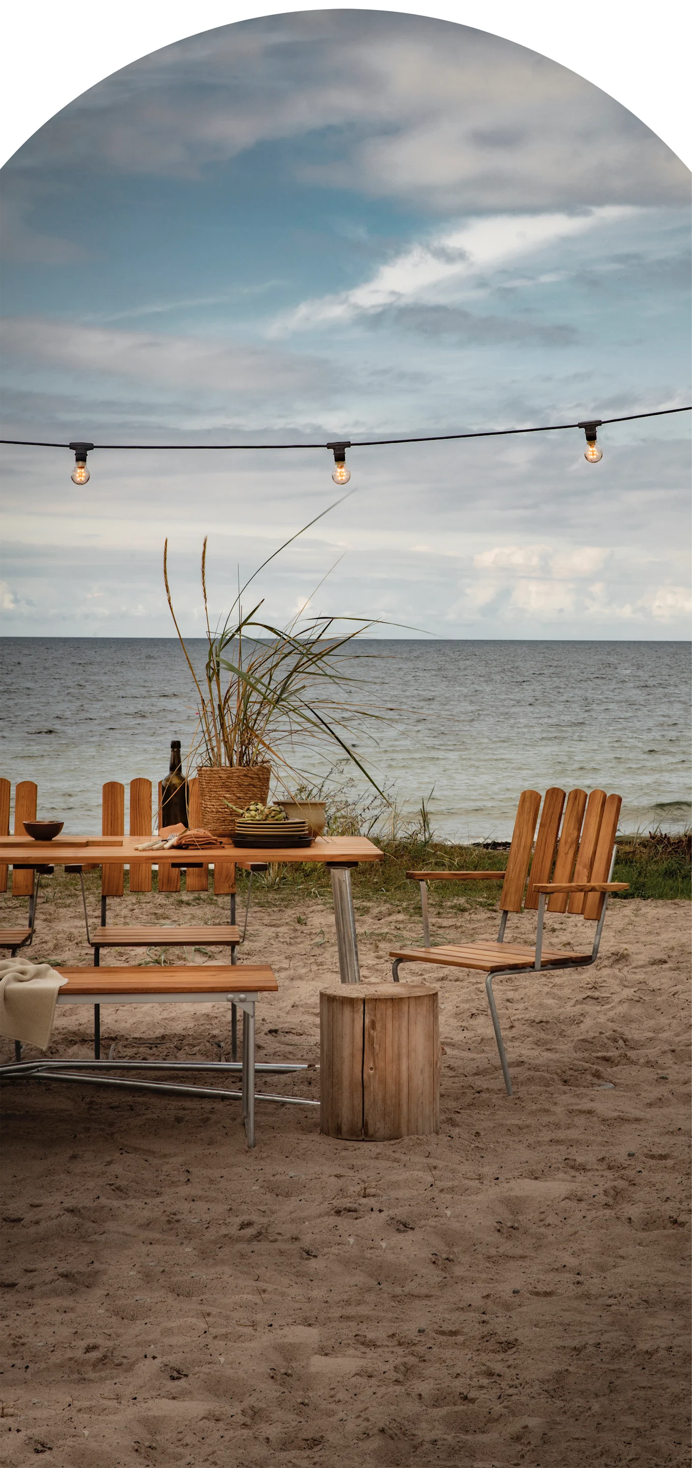 An outdoor wooden dining table and chairs on a sandy beach by the sea, with string lights overhead under a cloudy sky.