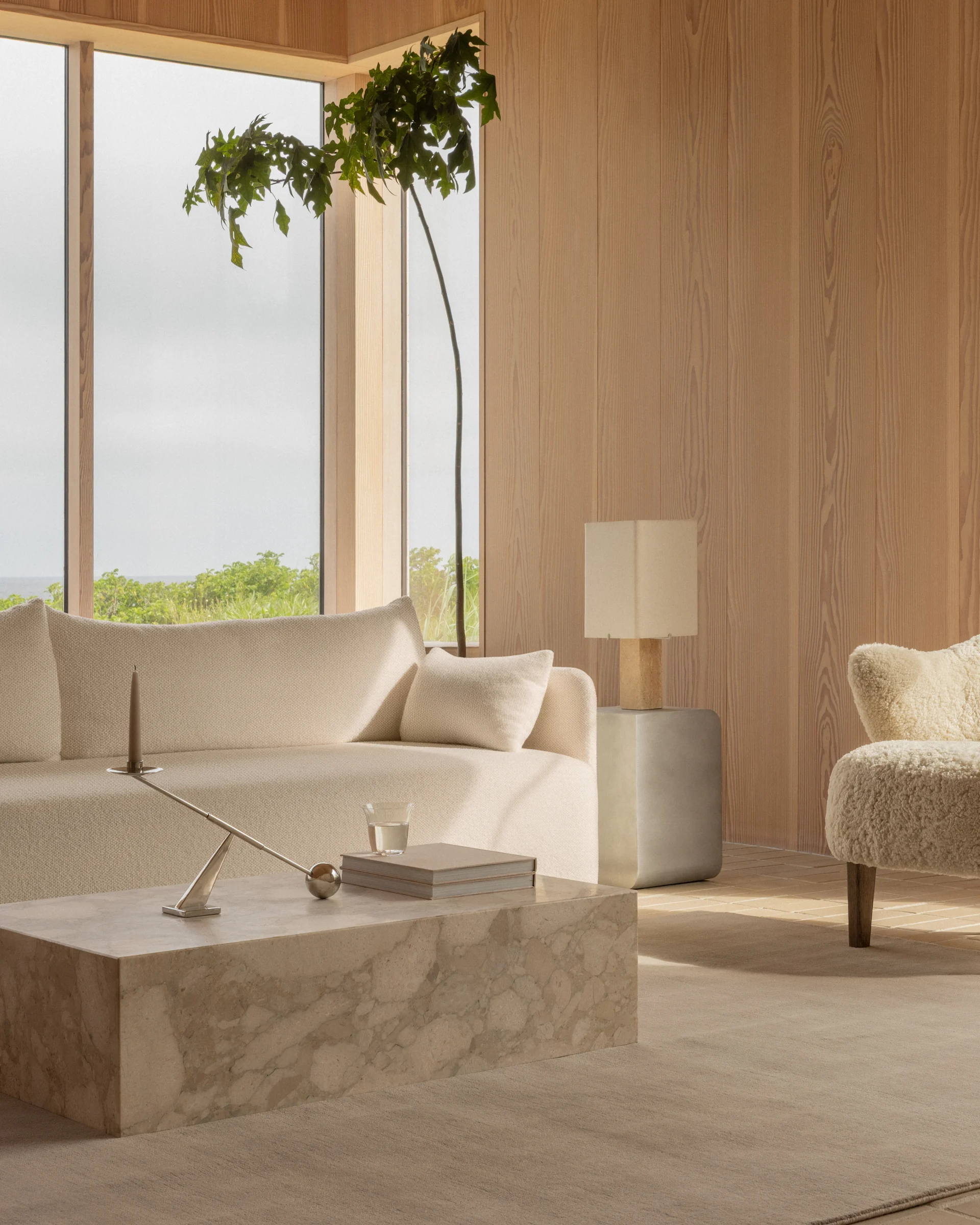 A serene living room with a cream textured sofa, marble coffee table, wooden walls, and large windows overlooking lush greenery.