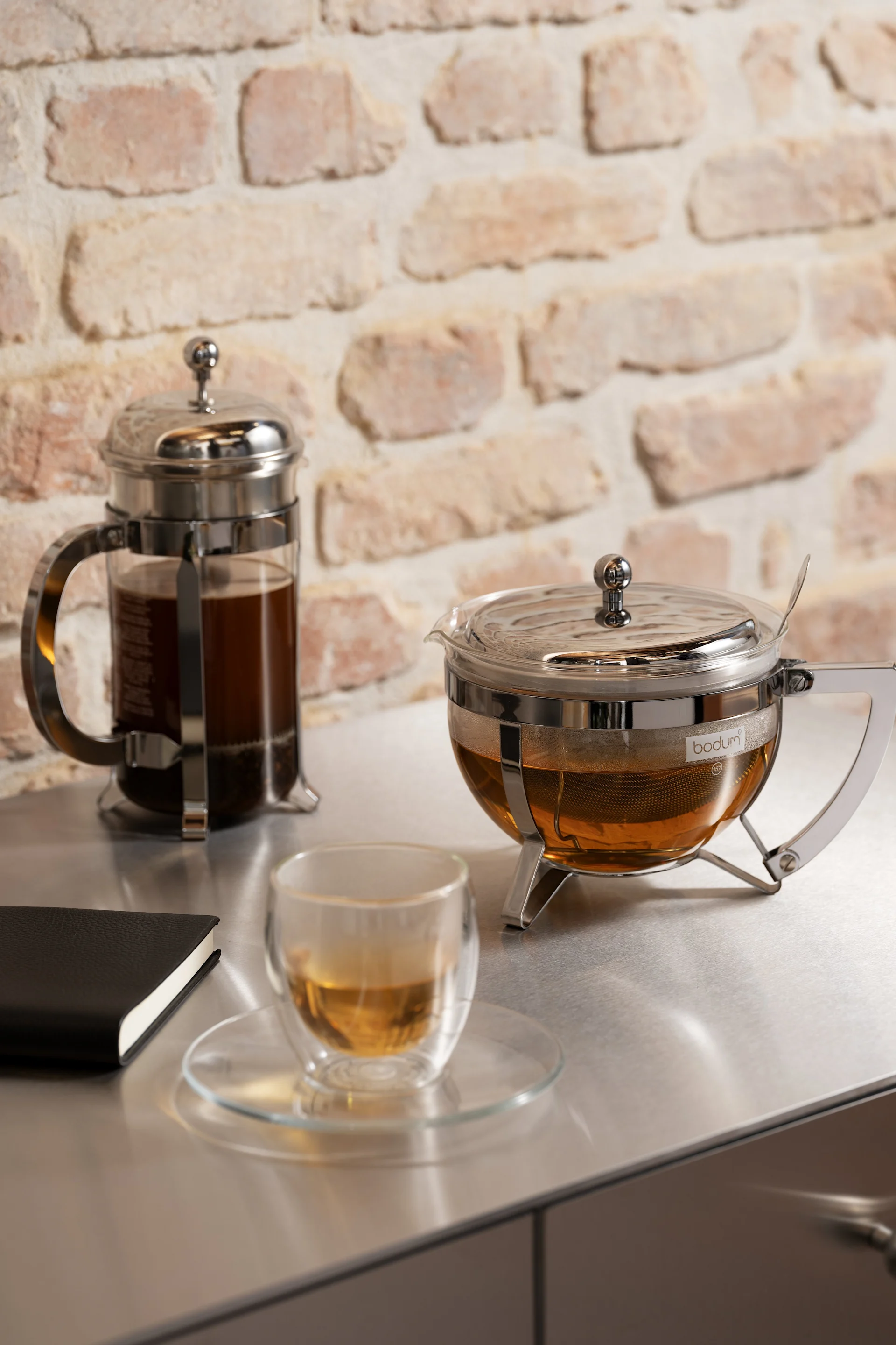 French press coffee maker, glass teapot with tea infuser, and teacup with amber tea on a metal counter with a brick wall.