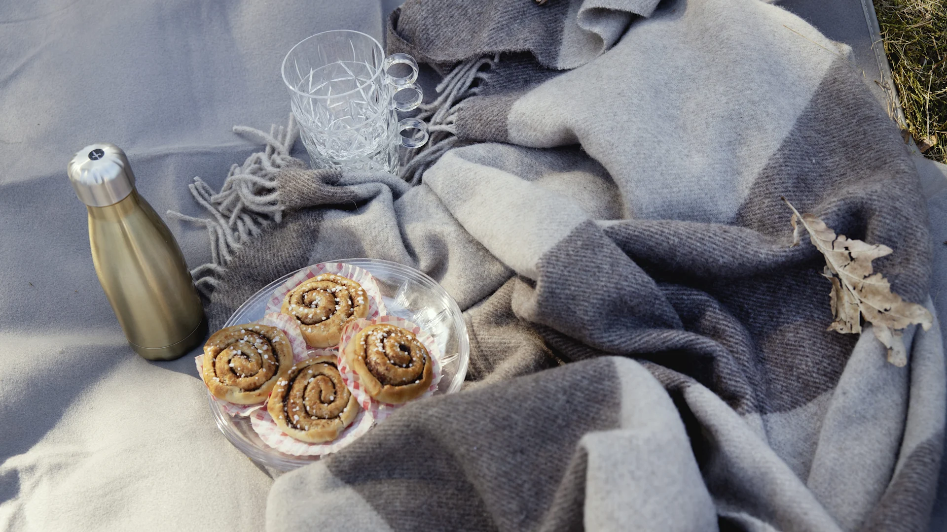 Picnic setup on a gray blanket with a golden thermos, two clear mugs, and a plate of cinnamon rolls.