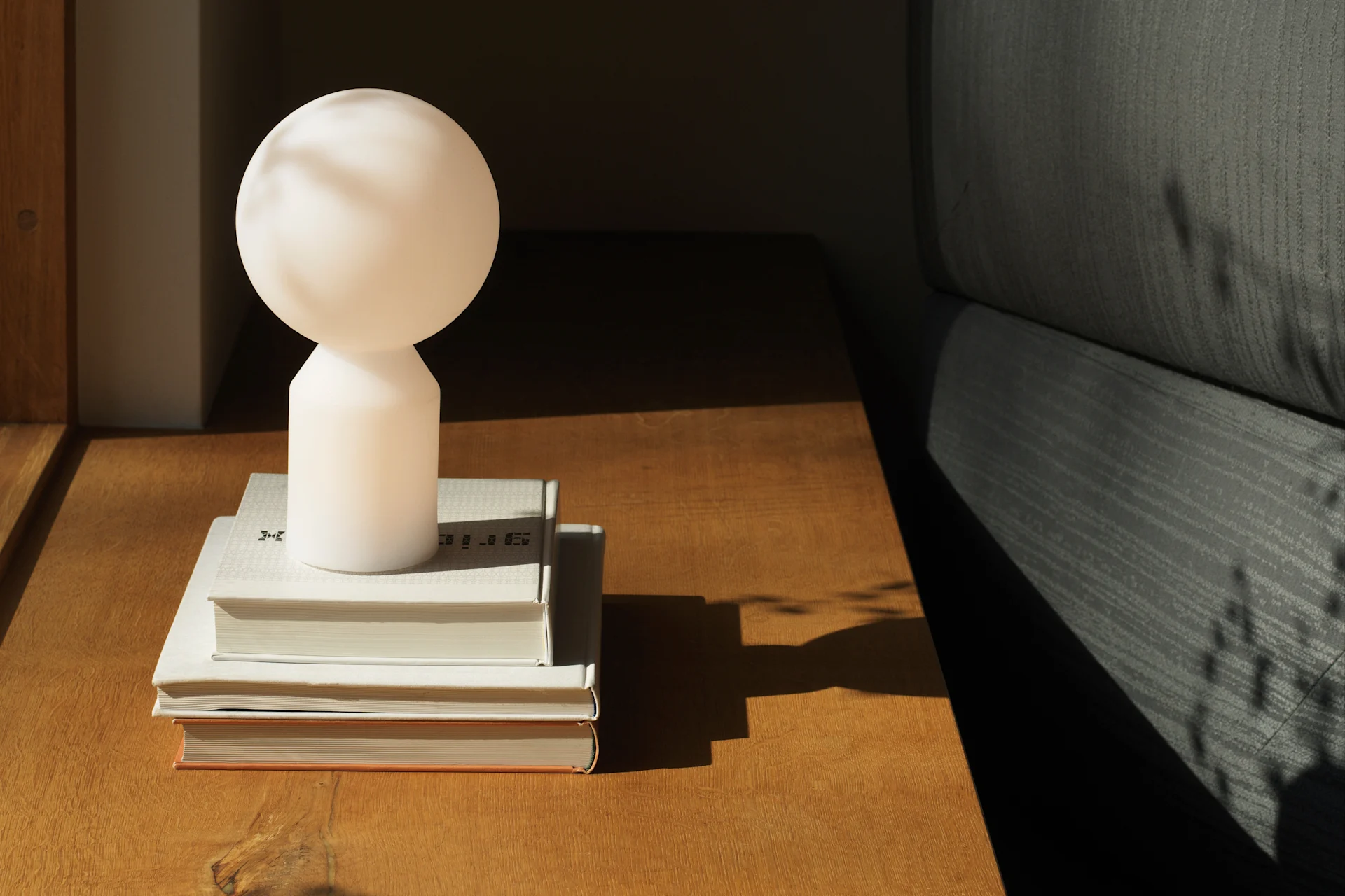 A spherical white lamp sits on a stack of books on a wooden table next to a gray sofa, with sunlight casting shadows.