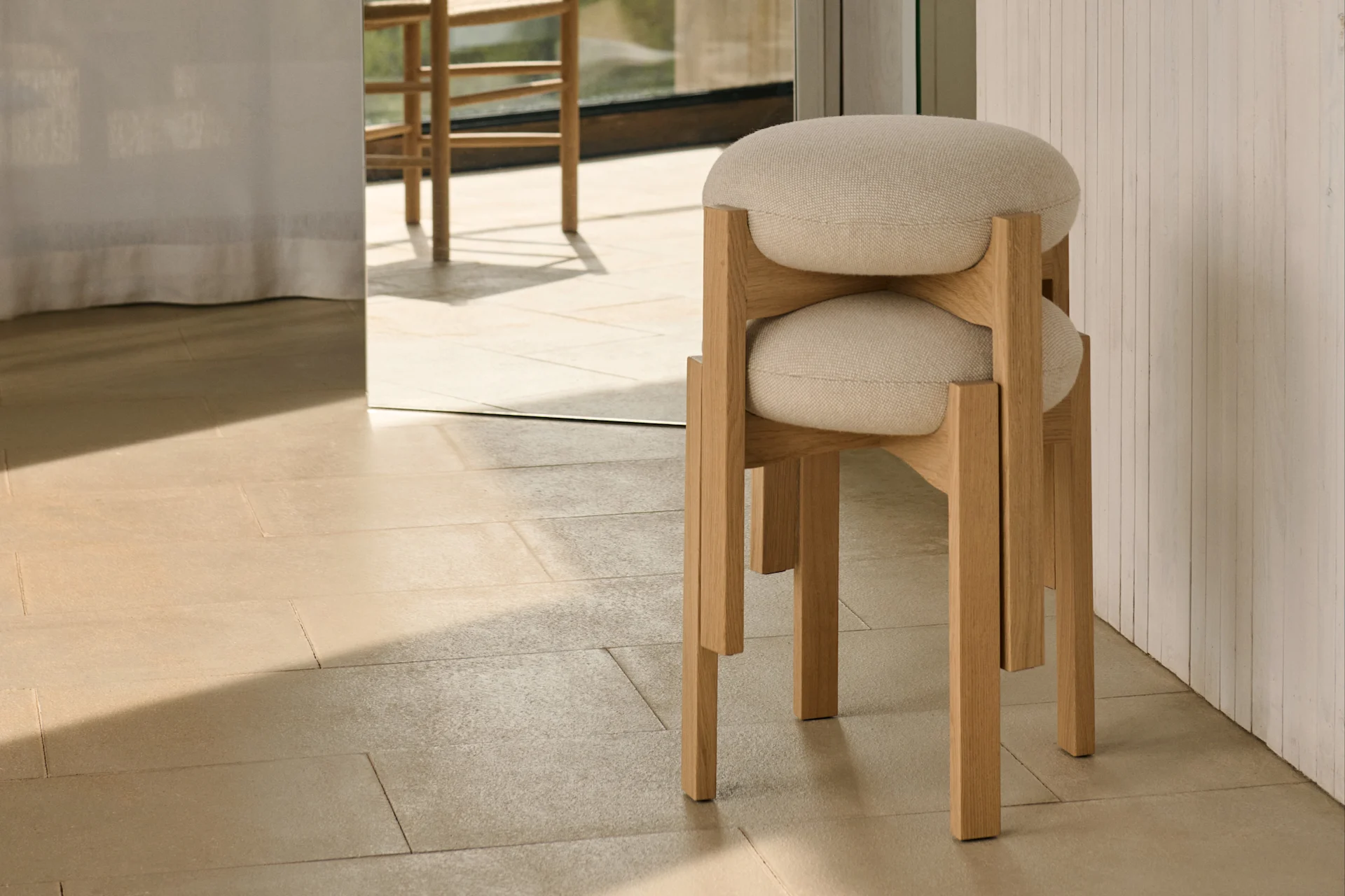 Two minimalist light wood stools with round upholstered seats are stacked on light tiles, with a sunny outdoor view through a large window.