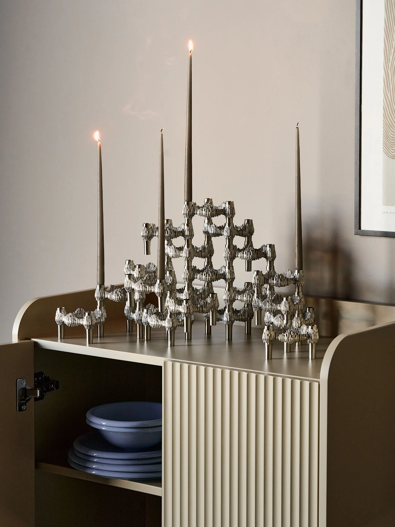 Sculptural silver candelabra with five lit grey candles on a light beige fluted cabinet.
