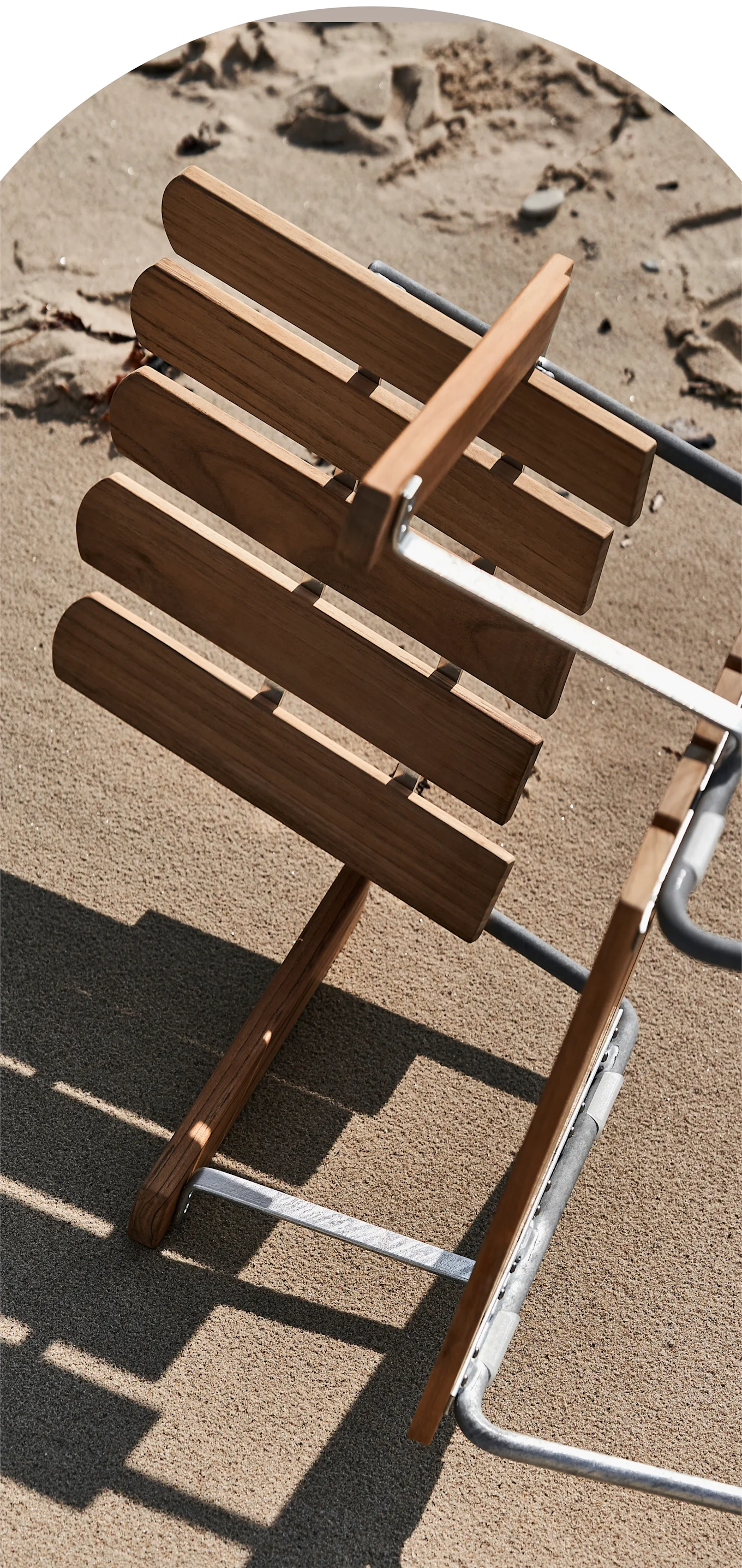 A close-up of a slatted wooden beach chair with a metal frame lying on light brown sand, casting shadows.