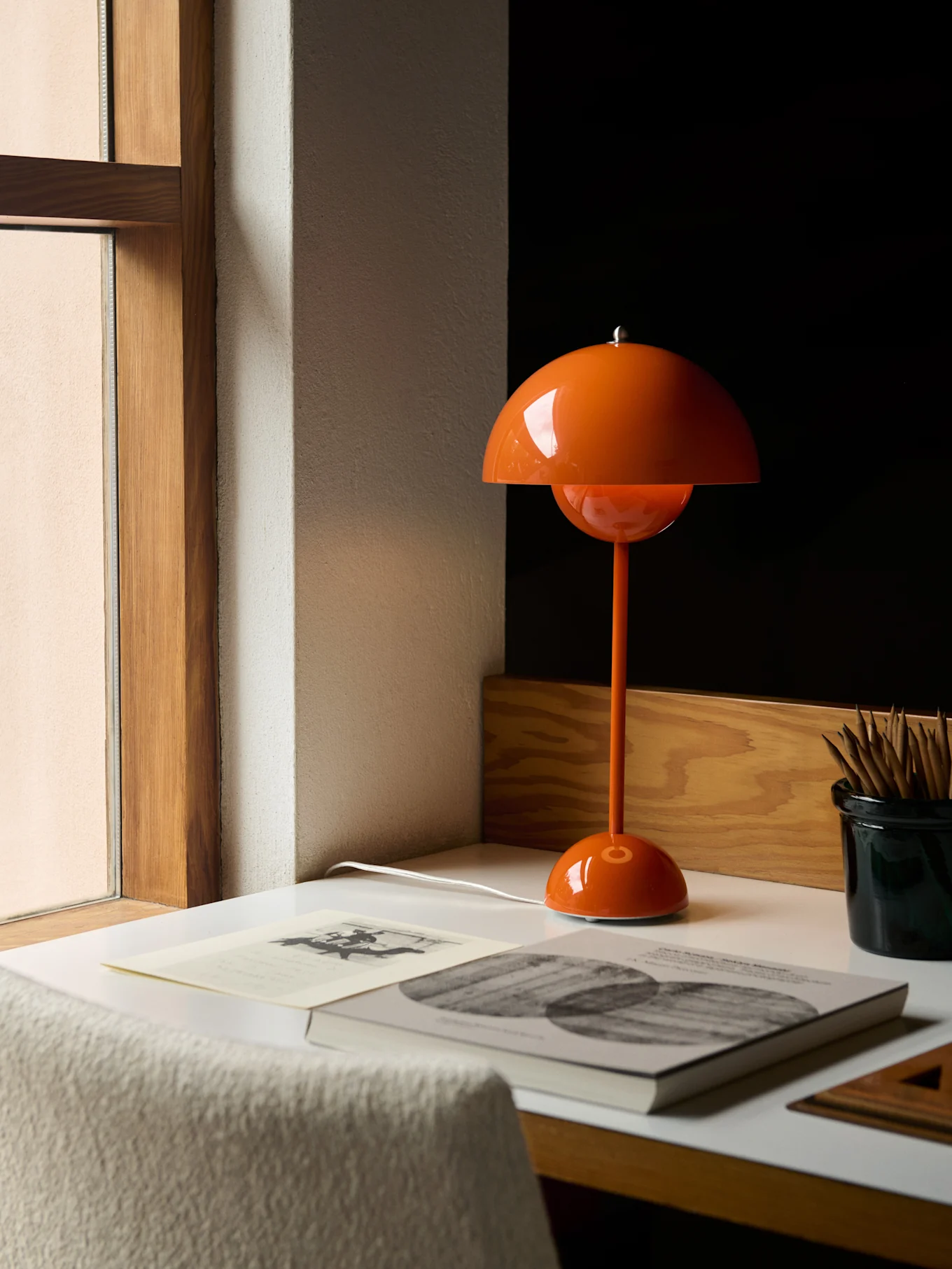 An orange mushroom-shaped table lamp on a white desk next to a window, with books and pencils.
