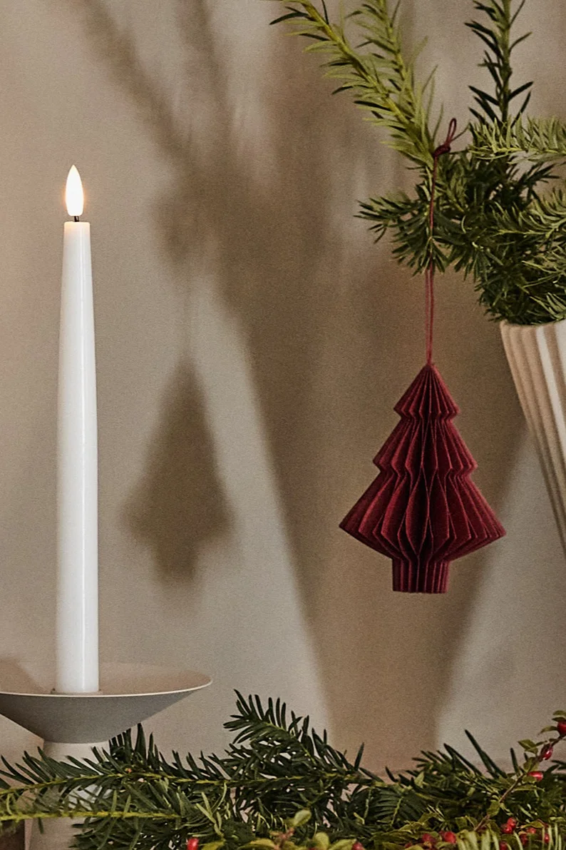 Lit white candle in a grey holder, a deep red folded paper Christmas tree ornament hangs from a green branch with berries.