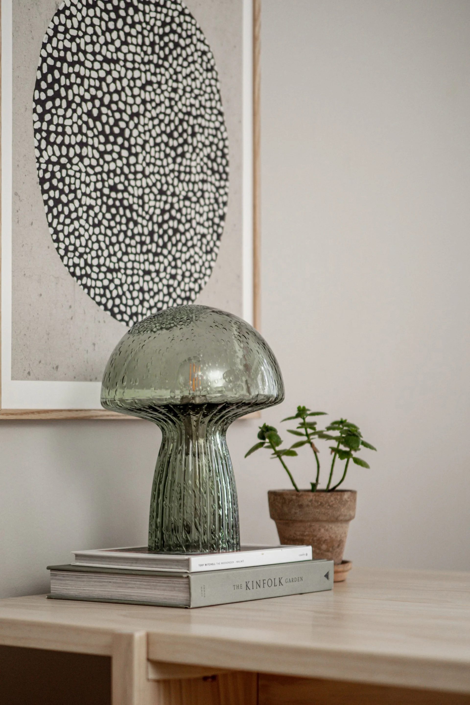 A green mushroom-shaped glass lamp on books next to a small plant, in front of an abstract dotted art print.
