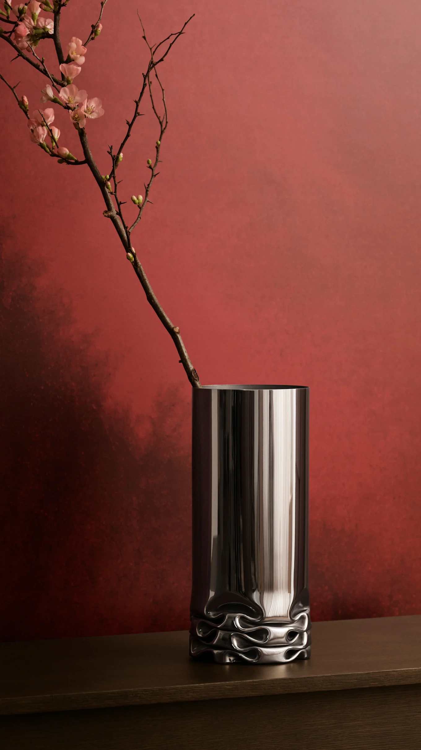 A tall dark silver vase with a wavy base holds a branch of light pink blossoms against a reddish-brown wall.