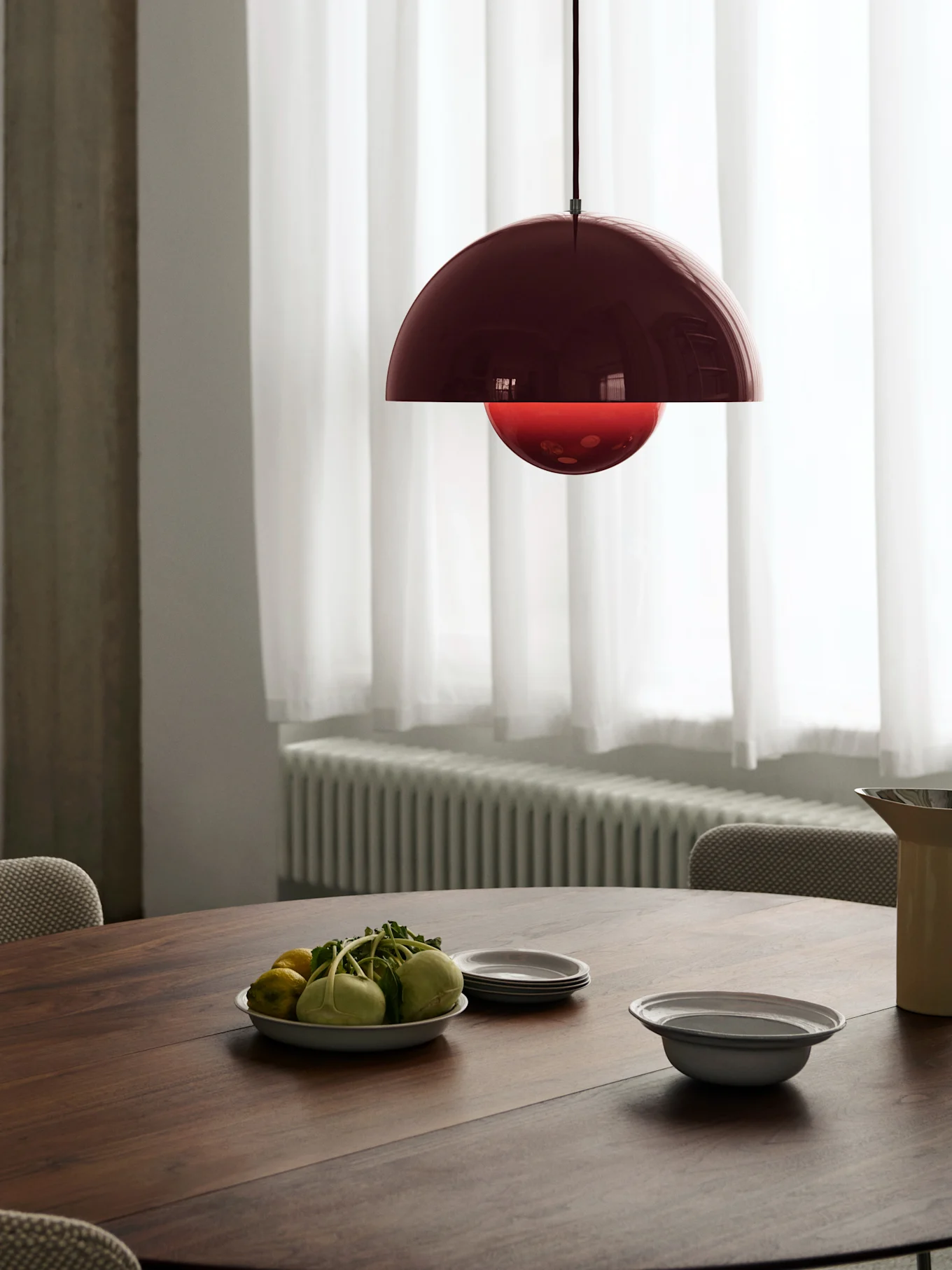 A dark red dome pendant lamp with a bright red inner globe hangs over a wooden dining table set with produce and plates.