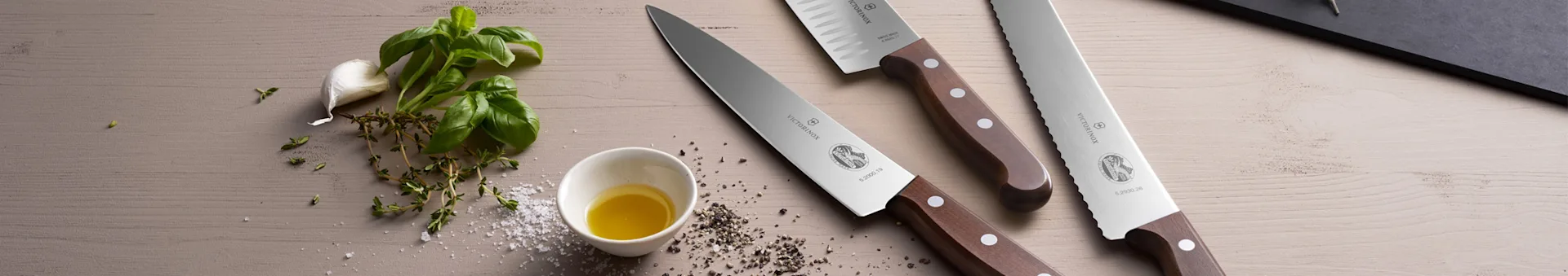 Three kitchen knives with wooden handles and various fresh ingredients on a light wooden surface.