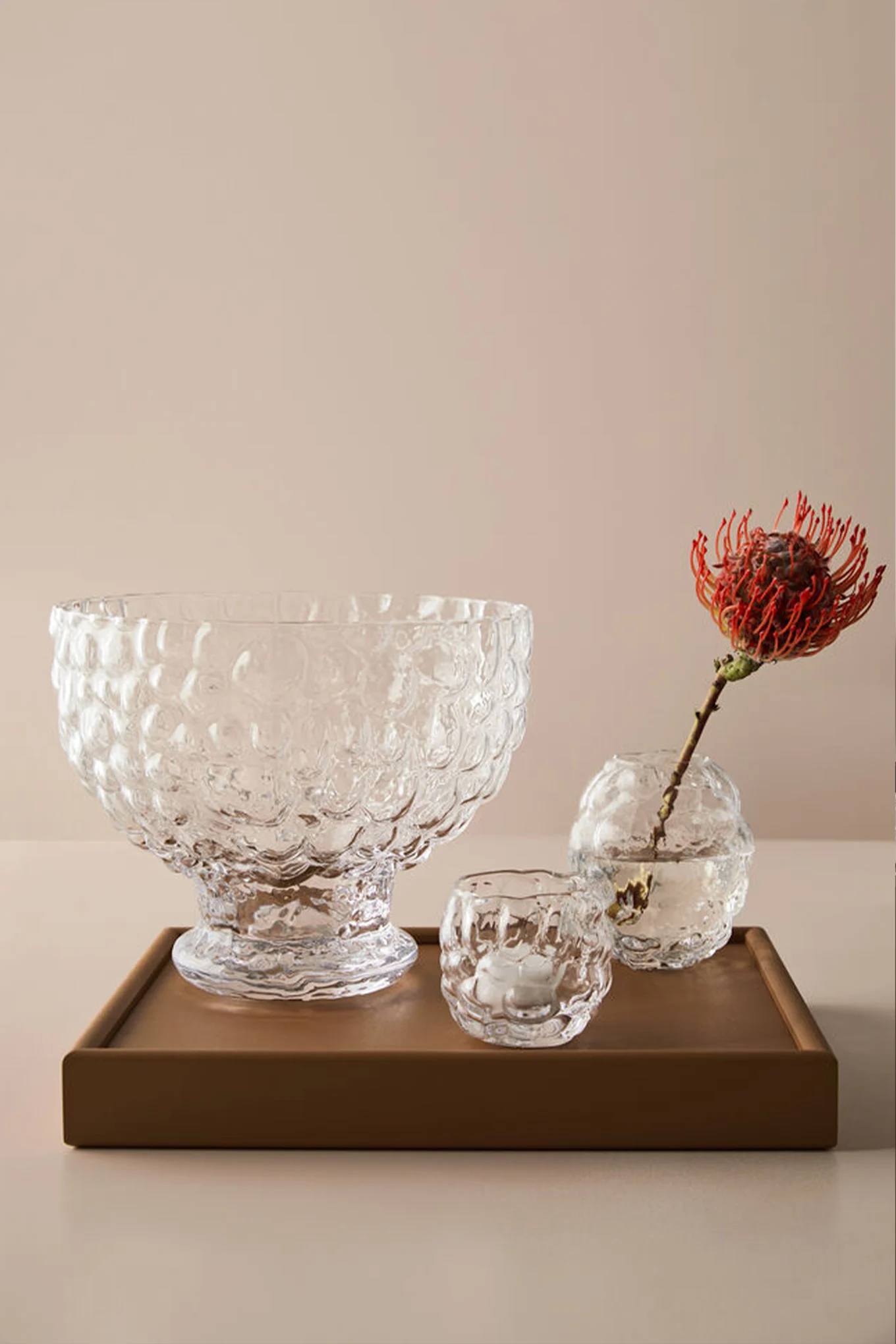 Three clear textured glass items on a brown tray: a large bowl, a votive with a candle, and a vase with a red protea flower.