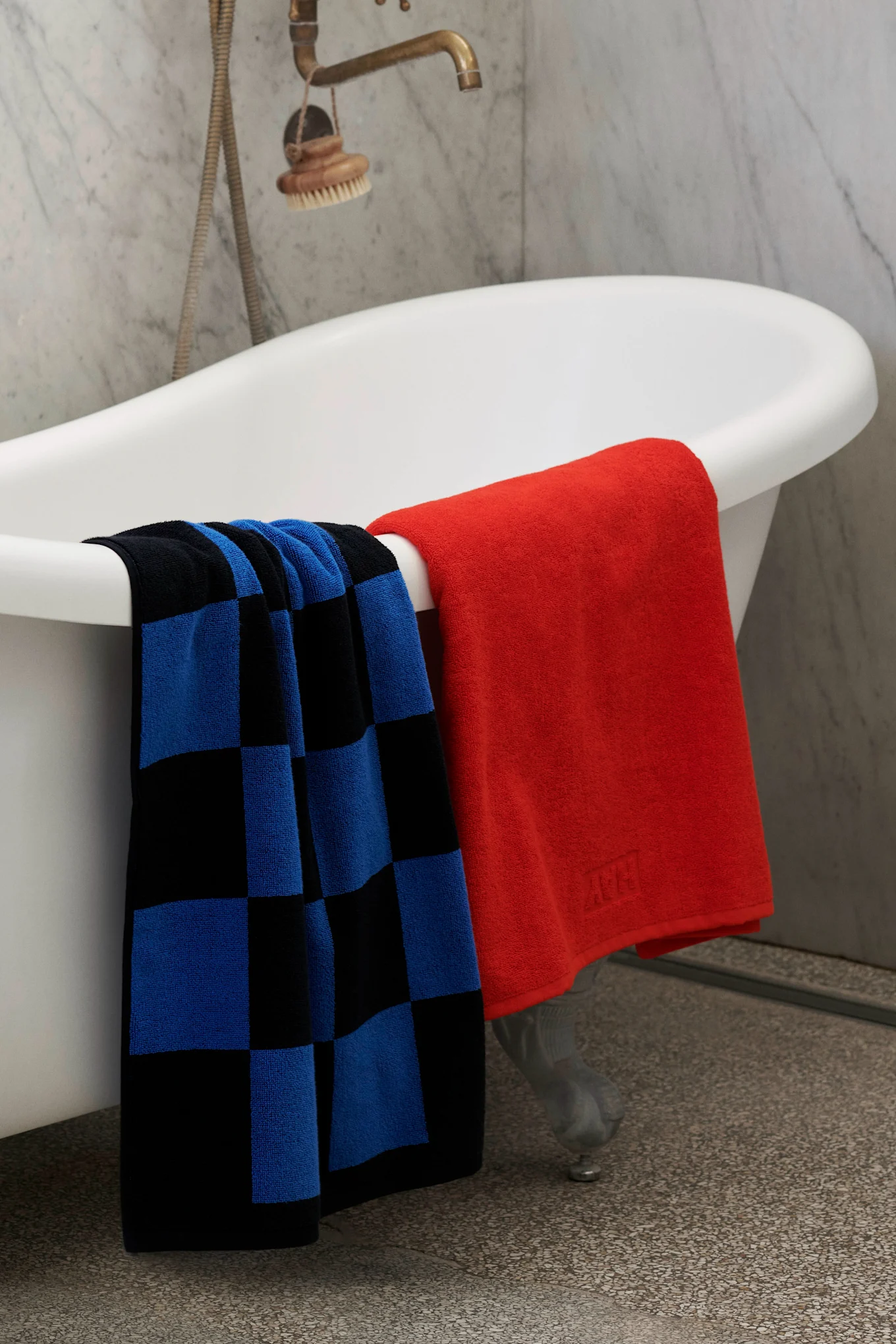 Two towels, one blue and black checkered and one solid red, draped over a white clawfoot bathtub in a bathroom.
