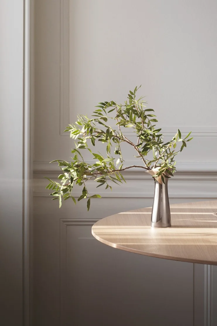 Silver vase with green leafy branches on a light wooden table, sunlight casting shadows on table and wall with molding.