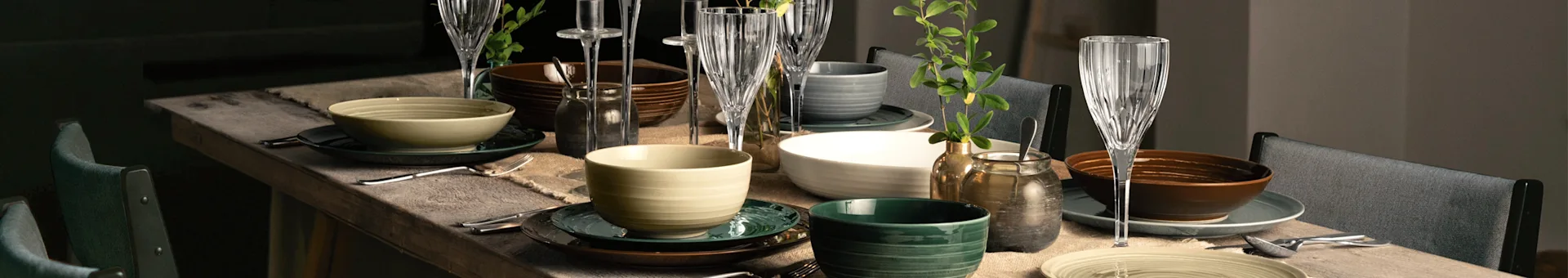 A dining table set with various bowls and plates in shades of green, brown, beige, and white, alongside clear glass goblets and plant decorations.