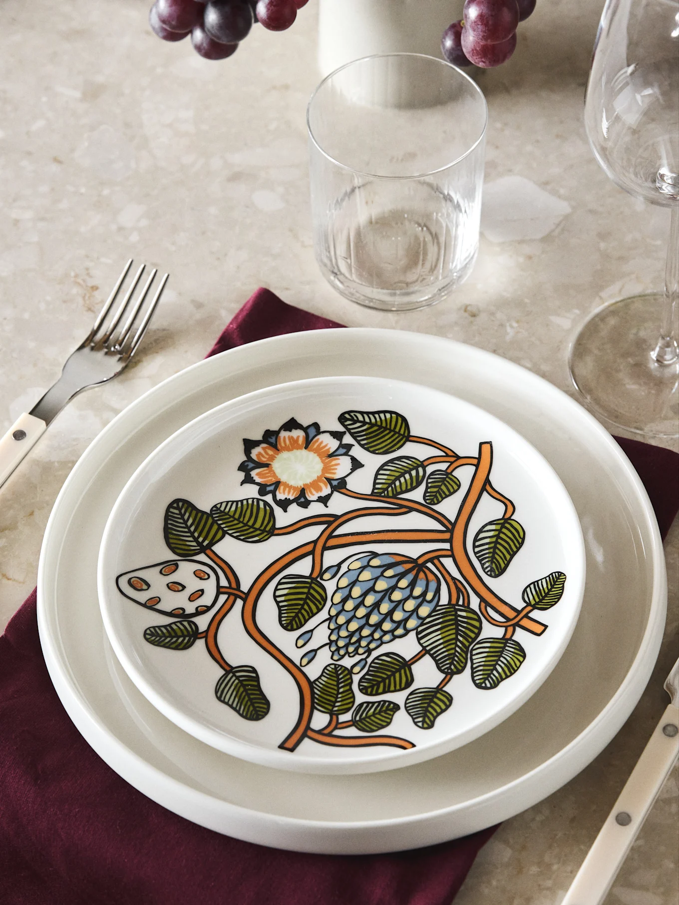 Two white plates, top one with a colorful floral and botanical design, on a red napkin with a fork.