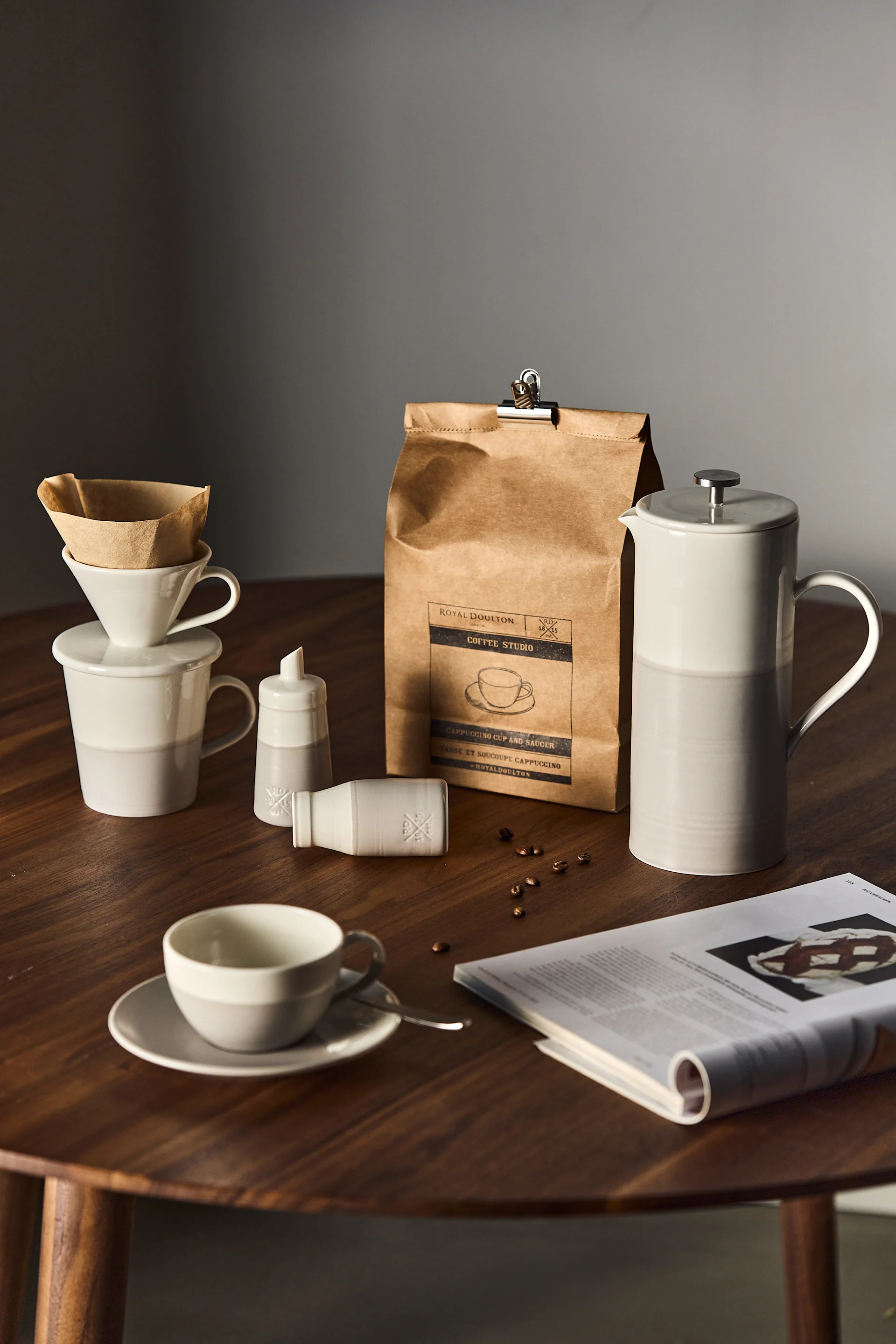 Ceramic white and grey coffee set with a French press, mugs, and a branded coffee bag on a wooden table.