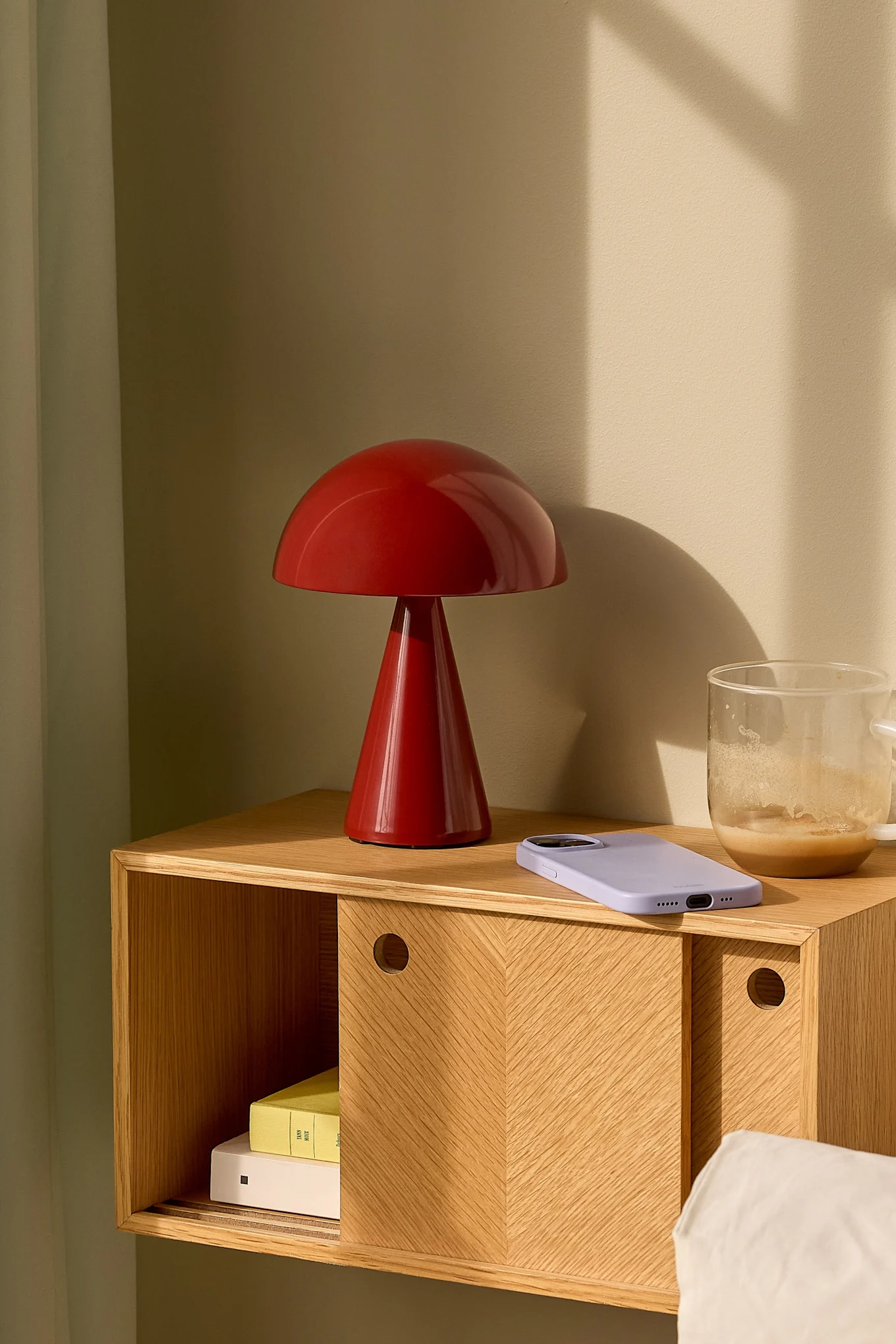 A red mushroom-shaped lamp, light purple smartphone, and glass coffee mug sit on a wooden nightstand with a sliding door.