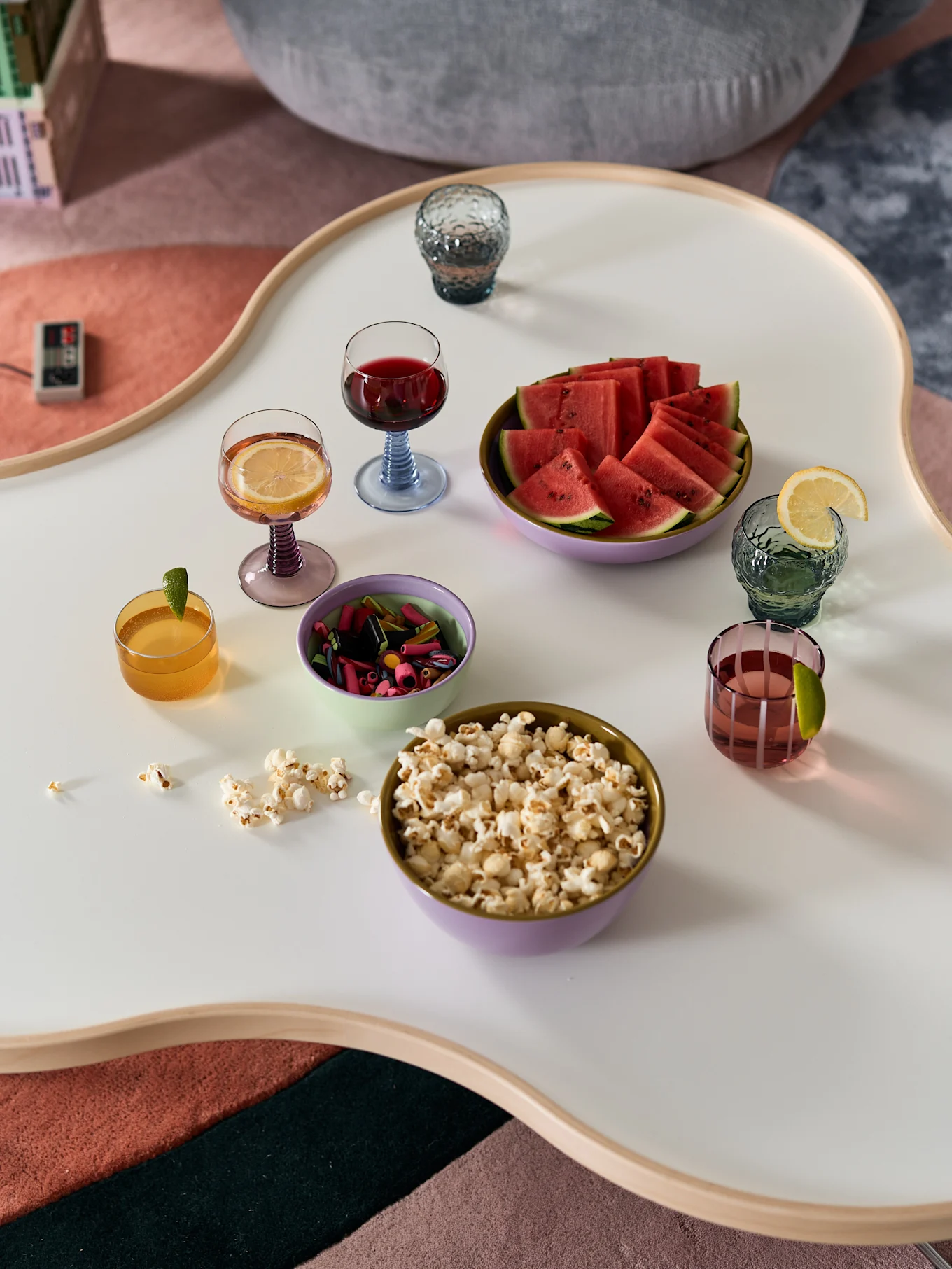 A white coffee table with popcorn, watermelon, candies, and five drinks. An old game controller is on a multi-colored rug.