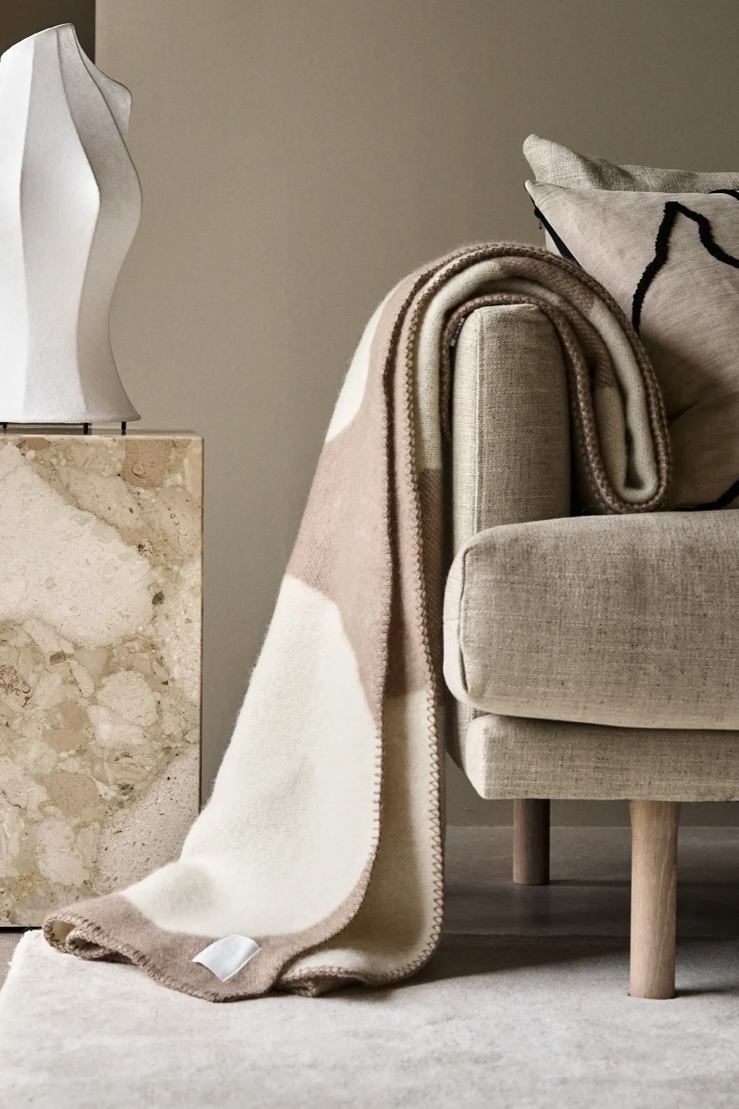 Beige sofa with a cream and taupe patterned throw blanket, next to a marble pedestal and white sculptural vase.