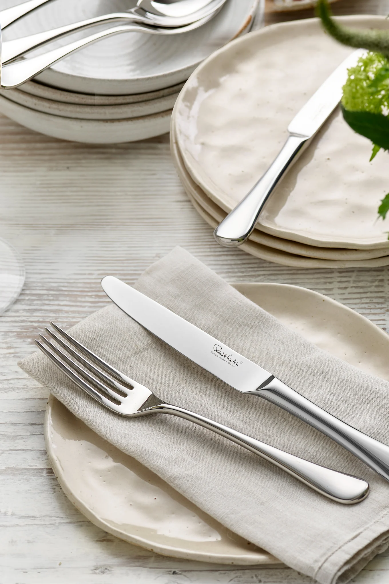 A close-up of a dinner table setting with a polished fork and knife on a linen napkin and rustic light-colored plates.