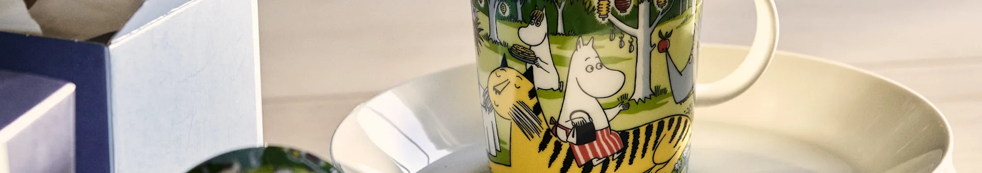 Moomin mug with Moomintroll holding pancakes and Snorkmaiden in a forest scene with lanterns, on a white saucer.