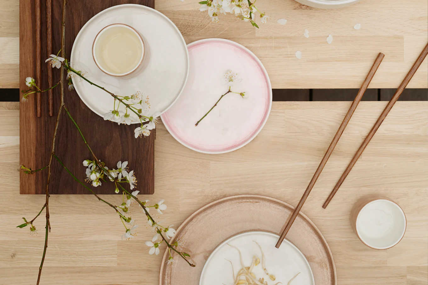 Serene table setting with neutral ceramic dishes, wooden chopsticks, tea, bean sprouts, and white blossoms on a light wooden surface.